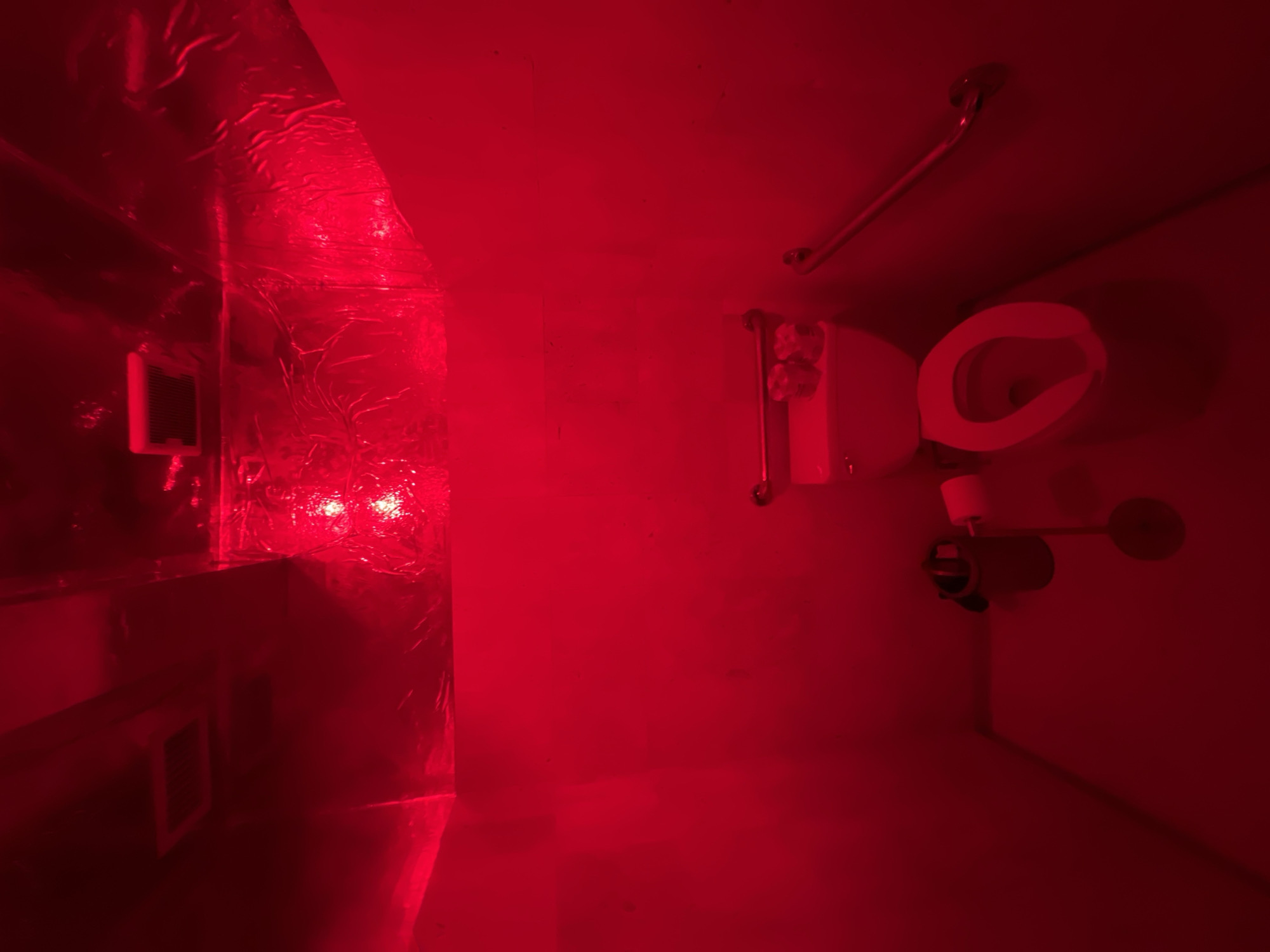 The image depicts an interior space, possibly a small bathroom, illuminated by a strong red light, likely from an emergency light fixture. On the right side of the frame, there is a toilet, equipped with a grab bar on the wall above it. A toilet paper dispenser is also present near the toilet. The left side of the image features some sort of duct or vent covered in a metallic, crinkled material. A small, rectangular grate or vent is also visible on the left wall. The source of the red light appears to be located near the center of the duct/vent, causing a bright reflection. The image is lacking in detail due to the overwhelming red light, making specific identification of materials and textures difficult. The location is undetermined. No people, actions or interactions are visible.