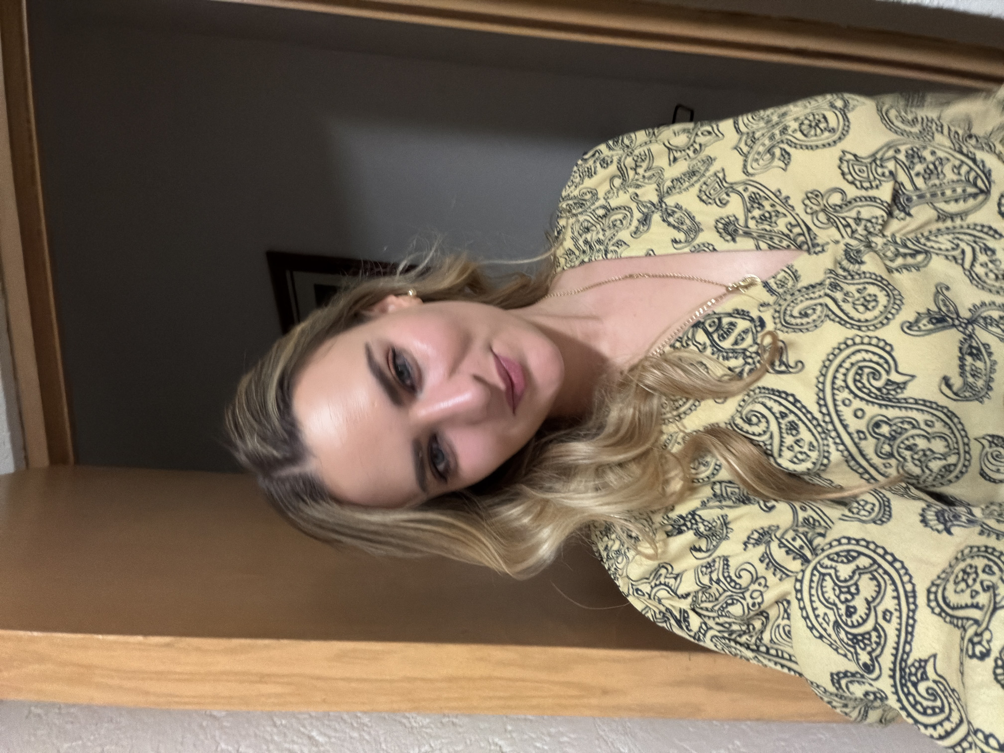 A woman with blonde hair and fair skin is wearing a yellow shirt with a black paisley pattern and a gold necklace. She is looking at the camera with a slight smile. The background is blurred, but there is a wooden frame visible, suggesting she may be standing in a doorway. There is a dark grey wall visible behind her and part of a framed picture is seen on the wall.
There is no information to indicate the image was taken in San Bernardino Tlaxcalancingo, Mexico.