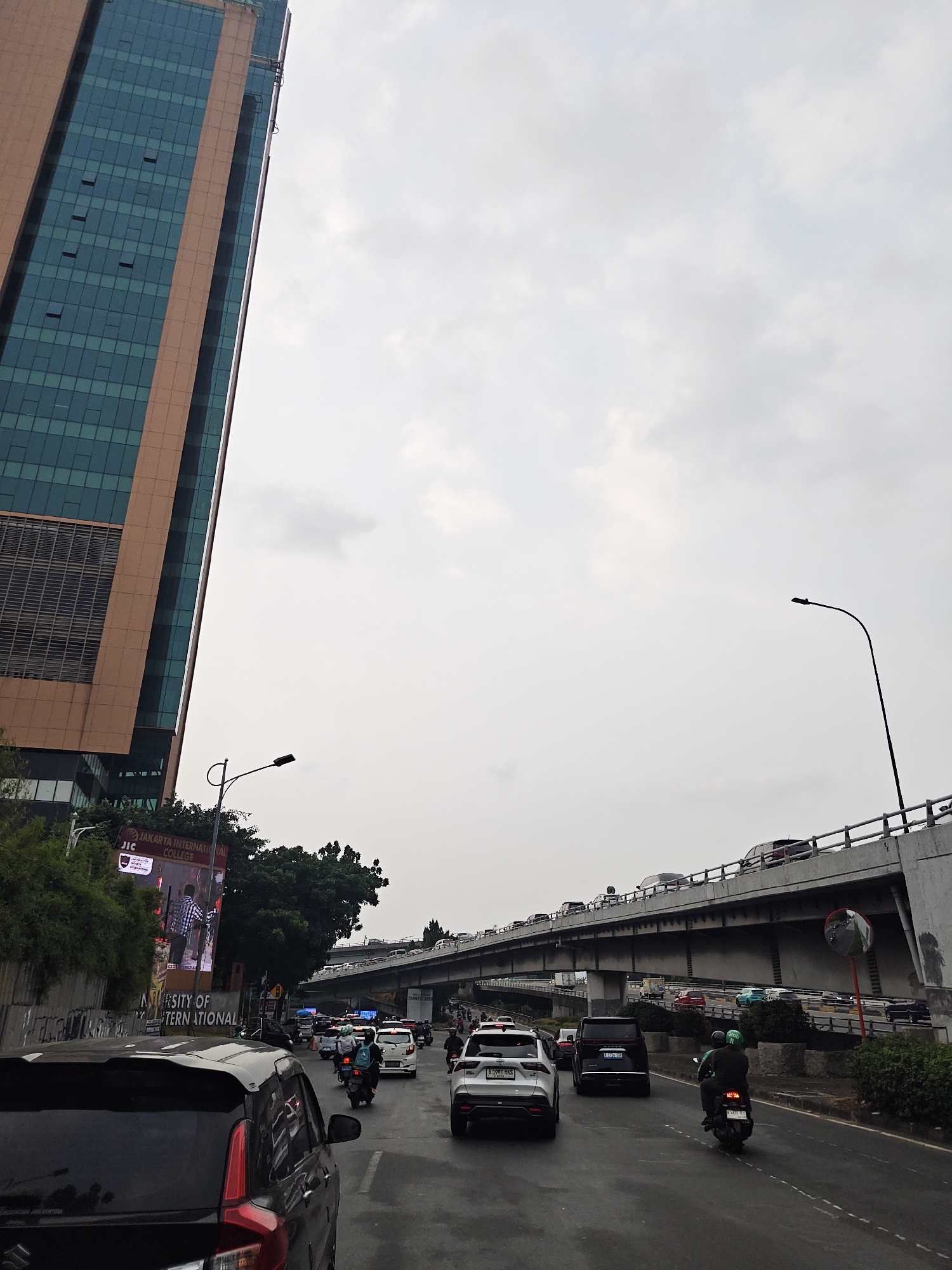 The image captures a street scene in West Jakarta, Indonesia, under a cloudy sky. In the foreground, a black car is seen from behind, with its taillights illuminated. To the right of the black car, a white SUV is visible, followed by a dark SUV. Further ahead, a motorcyclist wearing a green helmet is riding a motorcycle. In the background, a multi-level overpass is filled with traffic, with numerous cars and motorcycles visible. On the left side of the image, a tall modern building with a glass facade and an orange frame dominates the skyline. Below the building, trees and a billboard advertising "JAKARTA INTERNATIONAL COLLEGE" are present. The road appears wet, suggesting recent rain or high humidity. The overall scene depicts a busy urban environment with significant traffic congestion.