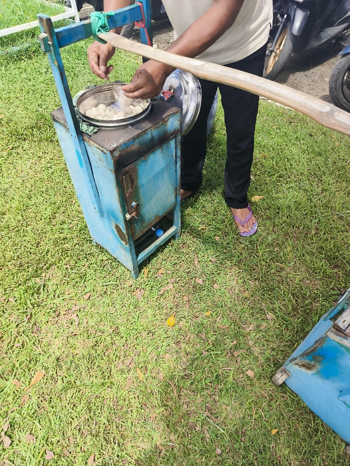 A man is preparing food on a street food cart in Majalengka, Indonesia. The cart is painted blue and has a metal shelf. The man is wearing a white shirt and dark pants and is using a spoon to stir food in a metal pot. He is standing on the ground, which is covered in green grass. The cart is positioned on a green lawn. The man is at a location in Majalengka with coordinates latitude: -6.846, longitude: 108.204. The cart is positioned next to an unknown object, likely another food cart, and is partly obscured from the view. The man is facing away from the viewer.