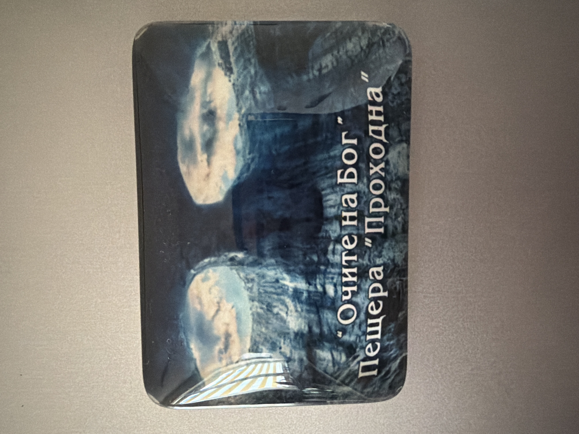 A rectangular magnet featuring a cave entrance with two openings resembling eyes and clouds above them is attached to a metallic surface. The text on the magnet reads "Очите на Бог" and "Пещера "Проходна"". The location in Pazardzhik, Bulgaria, is not discernible from the image itself.