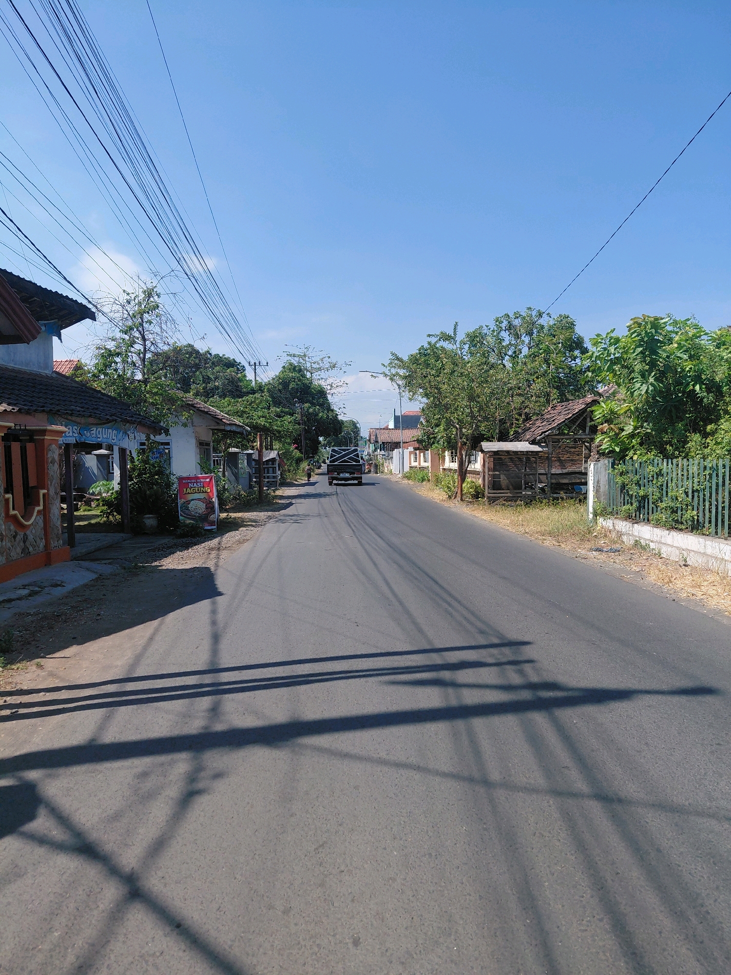 The image depicts a street scene in Jabung Sisir, Indonesia, under a clear blue sky. The asphalt road stretches into the distance, flanked by buildings and vegetation. On the left side of the road, there is a building with an orange and grey facade, a sign partially visible reading "MAS AGUNG," and a red advertising stand displaying an image of food. Further down the street on the left, more houses and trees are visible. On the right side of the road, a rustic wooden structure with a thatched roof stands next to a green metal fence. A black truck is driving away from the camera on the road, and a lone pedestrian is walking further down the street. A complex network of overhead power lines crisscrosses the sky. The ground along the roadside is covered with dry grass and some debris. The sun casts long shadows across the road, indicating it is likely daytime.