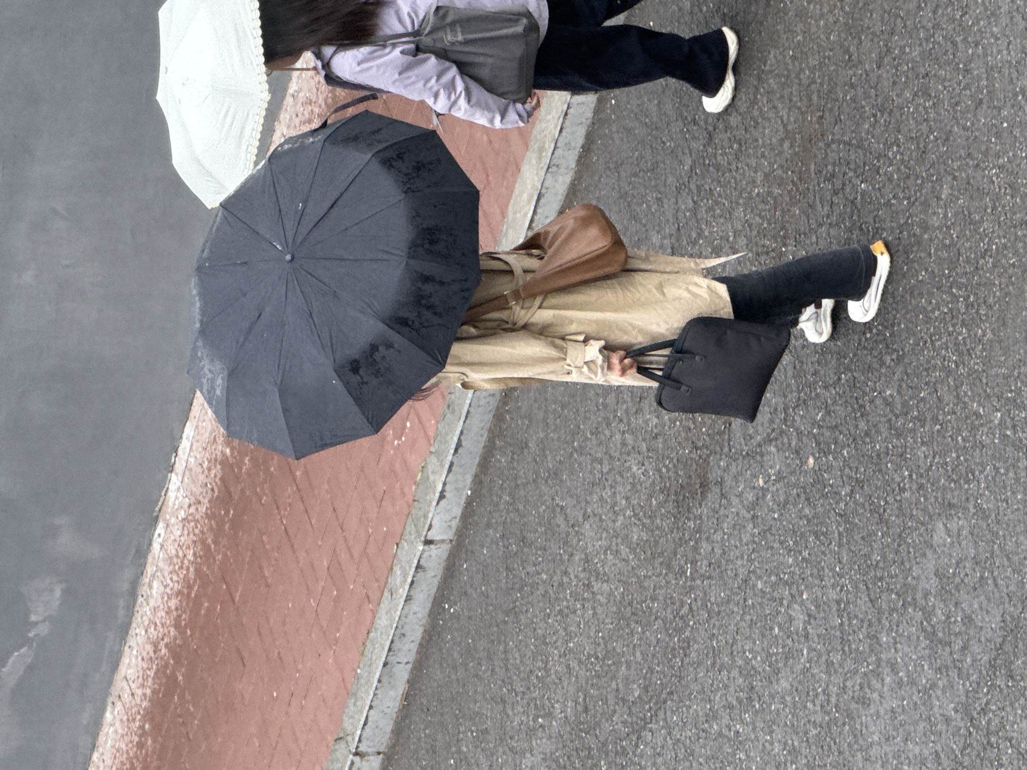 A high-angle shot depicts two pedestrians walking on a city street. The left pedestrian is holding a dark umbrella, wearing a beige coat, and black pants with white shoes. They are carrying a brown bag over their shoulder and a black bag in their hand. The right pedestrian has a light umbrella, wearing a light purple jacket and dark pants with white shoes. They are carrying a grey bag over their shoulder. The ground is a mix of wet pavement and red brick, suggesting it might be raining or have rained recently. The city is identified as Zhongtun, China.