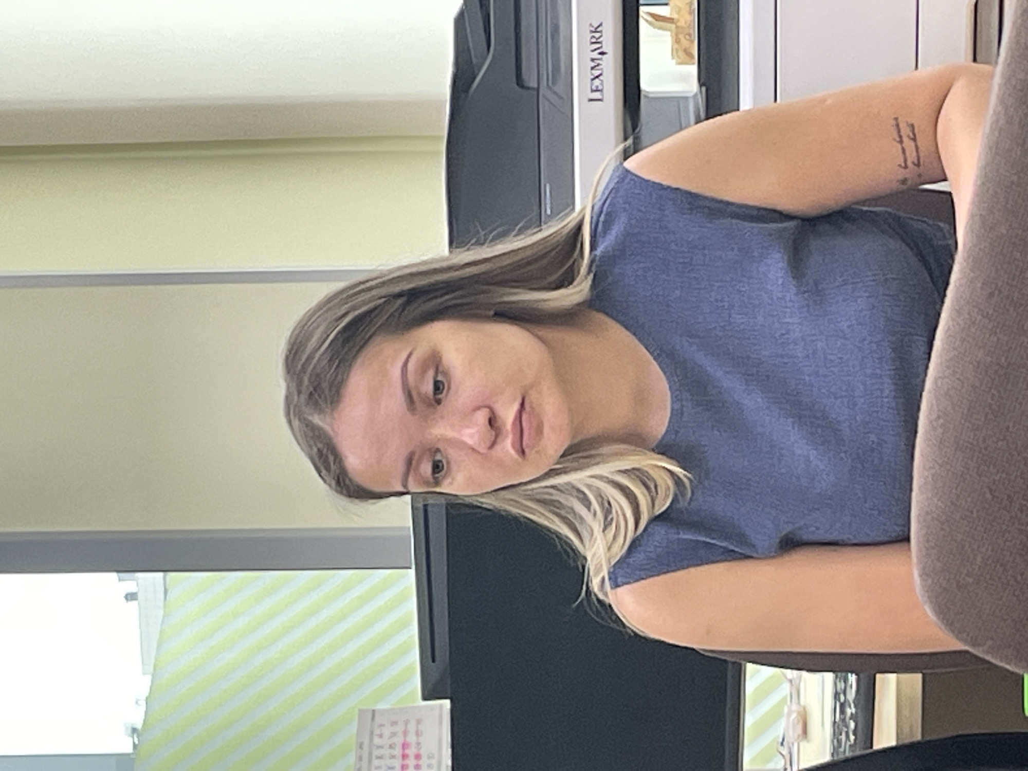 A head-and-shoulders portrait features a woman seated in an indoor office or workspace in Krasnoyarsk, Russia. She has long, straight hair with prominent blonde highlights and wears a plain blue short-sleeved top. Her head is slightly tilted, and she looks downwards with a neutral expression. A distinct tattoo composed of several vertical, stylized marks is visible on her left forearm.
The background includes a light green wall and a white ceiling. To the woman's right, a large, dark Lexmark multifunction printer or copier stands, with the brand name "LEXMARK" clearly visible in dark letters on a white panel. To her left, a portion of a window is visible, covered by light green and white striped blinds or curtains. Part of a desk calendar, displaying a grid with some unreadable text, is on a surface below the window. A brown chair armrest is visible on the far right of the frame, suggesting she is seated. The lighting indicates a daytime setting with ambient indoor illumination. The scene conveys a typical office environment.