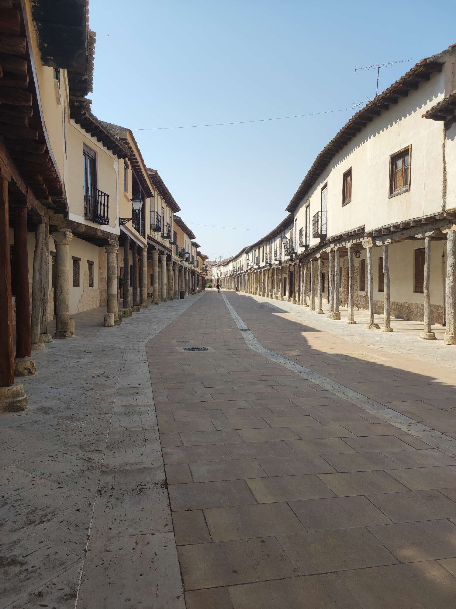 This is a vertical, eye-level, medium shot of a street in Ampudia, Spain. The street is lined on both sides by a long arcade of buildings. The buildings have cream-colored walls, red tile roofs, and wooden beams supporting overhangs. The arcades are supported by stone columns and wooden posts. The street is paved with large, flat stones. A single person is walking in the distance, toward the left side of the frame. The sky is clear and blue. The image appears to have been taken on a sunny day.