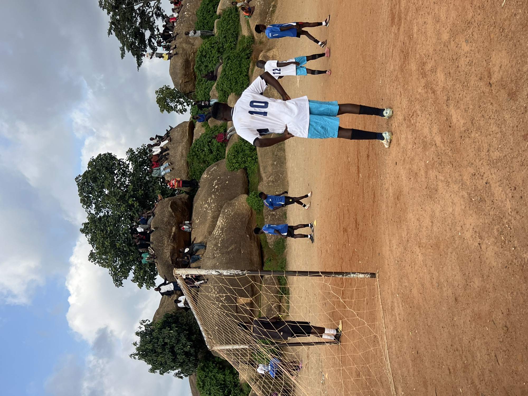 This is an outdoor, eye-level, full shot of a soccer game being played on a dirt field in Jos, Nigeria. In the foreground, a player wearing a white jersey with the number 10 and blue shorts stands with his hands on his hips. Behind him, players in blue jerseys and shorts are scattered across the field. A soccer net is visible on the left side of the frame. In the background, people are sitting on large rock formations, watching the game. Trees and greenery surround the rocks. The sky is cloudy. The ground is a reddish-brown dirt, characteristic of many parts of Jos, Nigeria.