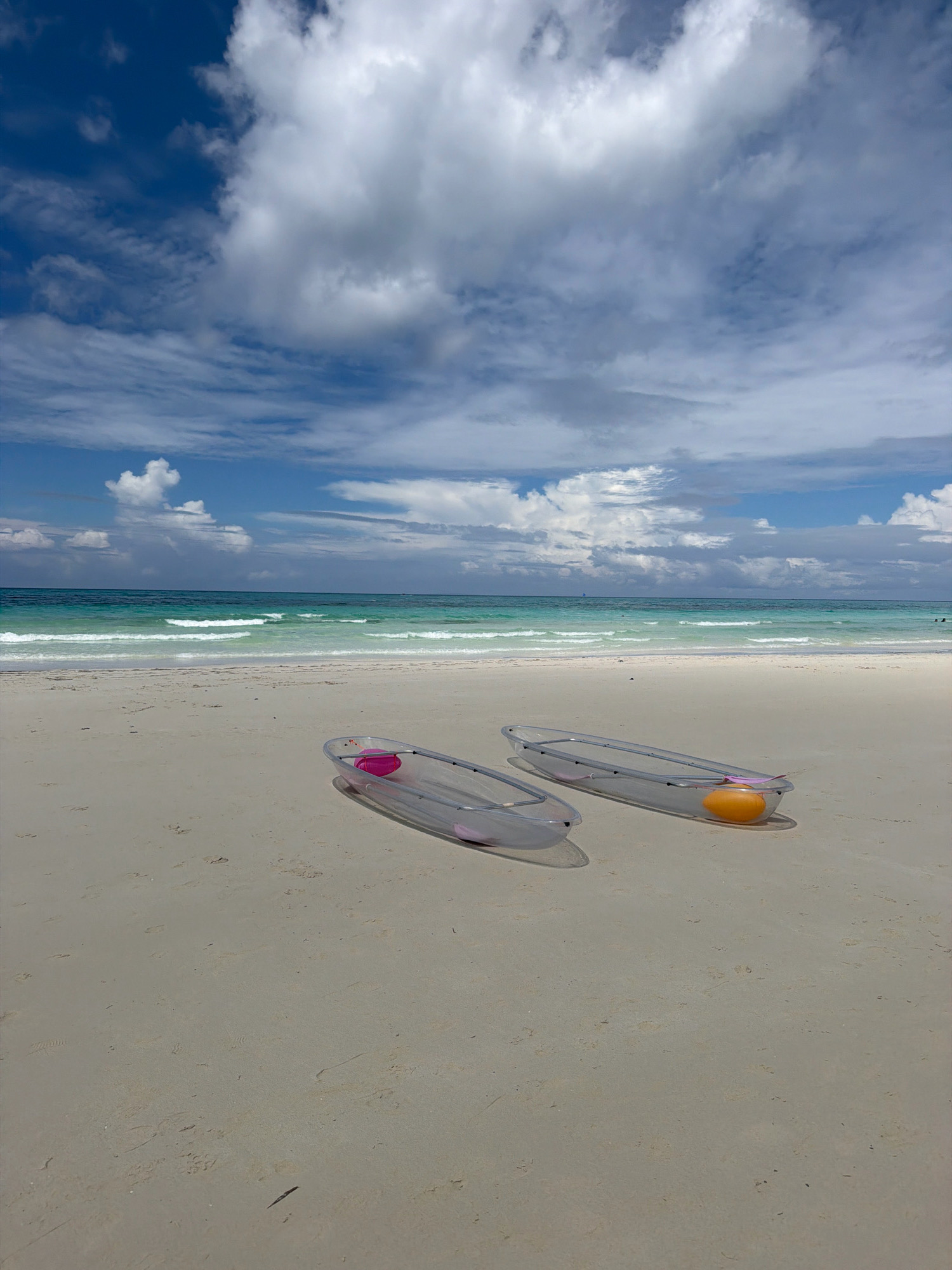 Two clear kayaks are positioned on a sandy beach. One kayak, closer to the foreground, contains a pink flotation device. The second kayak, further back, holds an orange flotation device. The beach is relatively empty, with subtle footprints visible in the sand. The ocean stretches out in the background, with gentle waves rolling towards the shore. The water transitions from a turquoise hue closer to the beach to a deeper blue further out. Above the ocean, a bright blue sky is dotted with numerous white, puffy clouds. The scene appears to be located at a beach in Malay, Philippines, likely Boracay, given the characteristic white sand and clear turquoise waters often associated with the area.