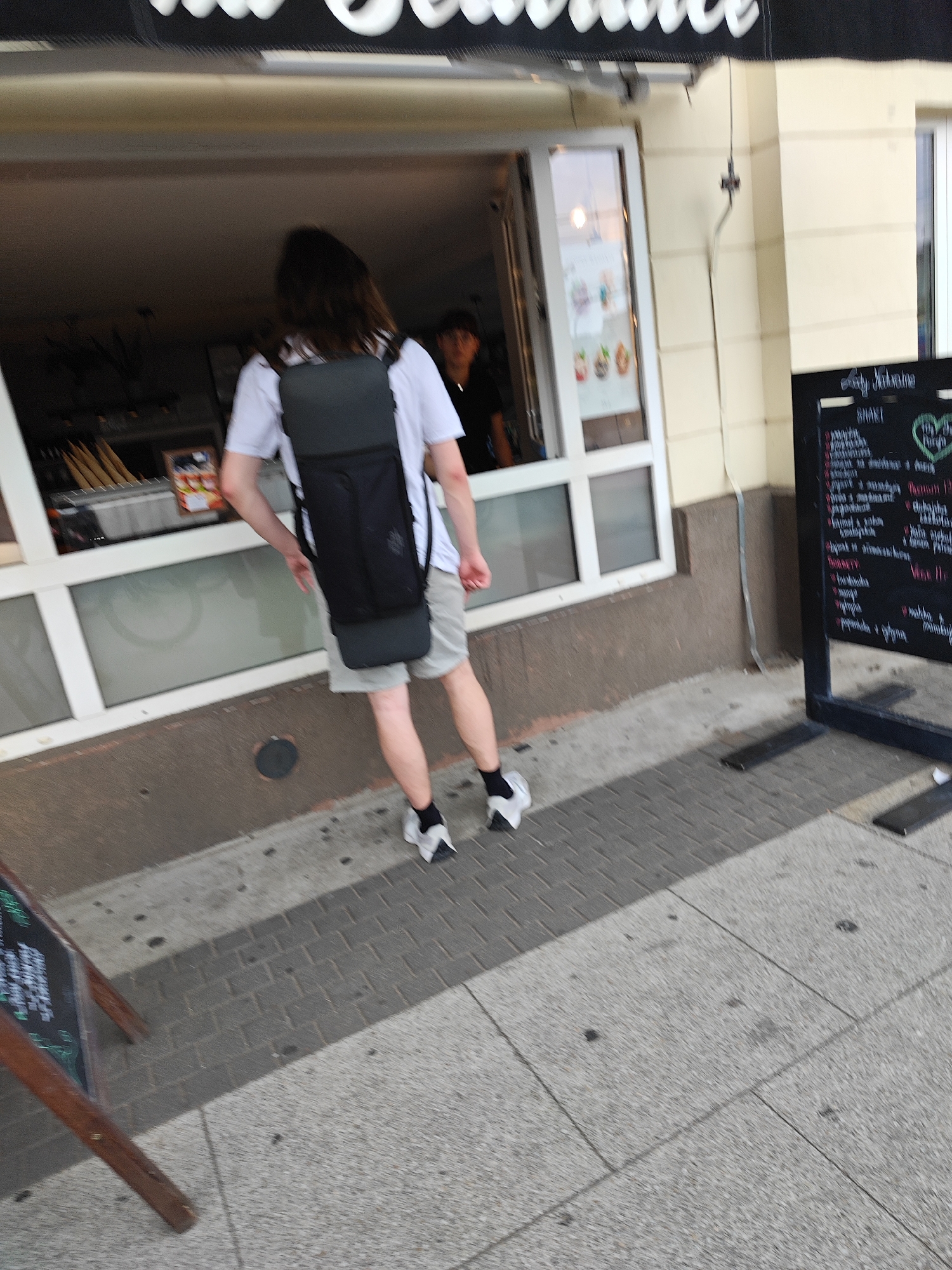 The image shows a person from behind, wearing a white t-shirt and light-colored shorts, with a black backpack. The person is walking on a sidewalk in front of a building with a window, possibly a shop or cafe. A black chalkboard menu stand is visible to the right of the person. The sidewalk is paved with grey bricks and concrete. In the background, through the window, another person can be seen. The overall lighting suggests it is daytime. The sign above the window, partially visible, has white lettering on a black background. This scene is located on the Stary Rynek (Old Market Square) in Poznan, Poland, a central square known for its historic buildings and bustling atmosphere.