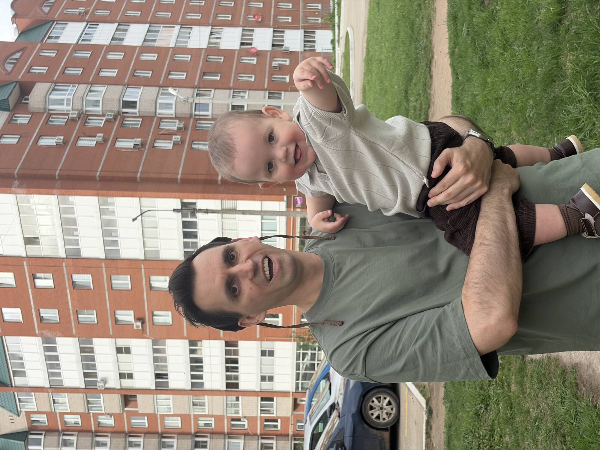 A man is holding a toddler. The man is wearing a green t-shirt. The toddler is wearing a light-colored shirt and brown shorts and shoes. The man is smiling and looking at the camera. The toddler is also smiling and has his arm raised. In the background, there is a large apartment building with many windows and balconies, a blue car, grass, and trees. The location is Chaikovsky, Russia, based on architectural style.
