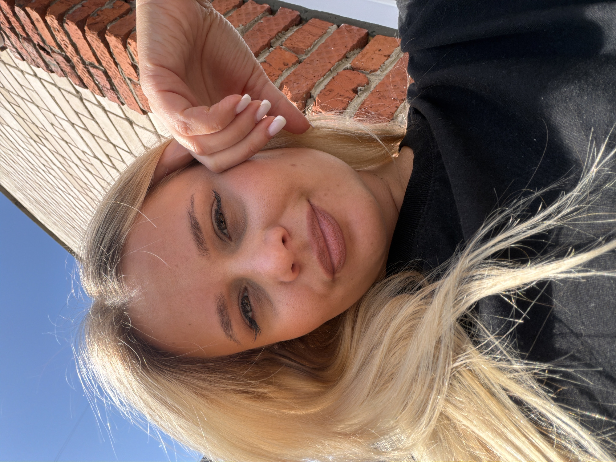 A blonde woman is seen in an upside-down selfie with a brick building and blue sky in the background. The woman is wearing a black garment, and her hand is near her forehead. The photograph appears to have been taken outdoors in Sarapul, Russia.