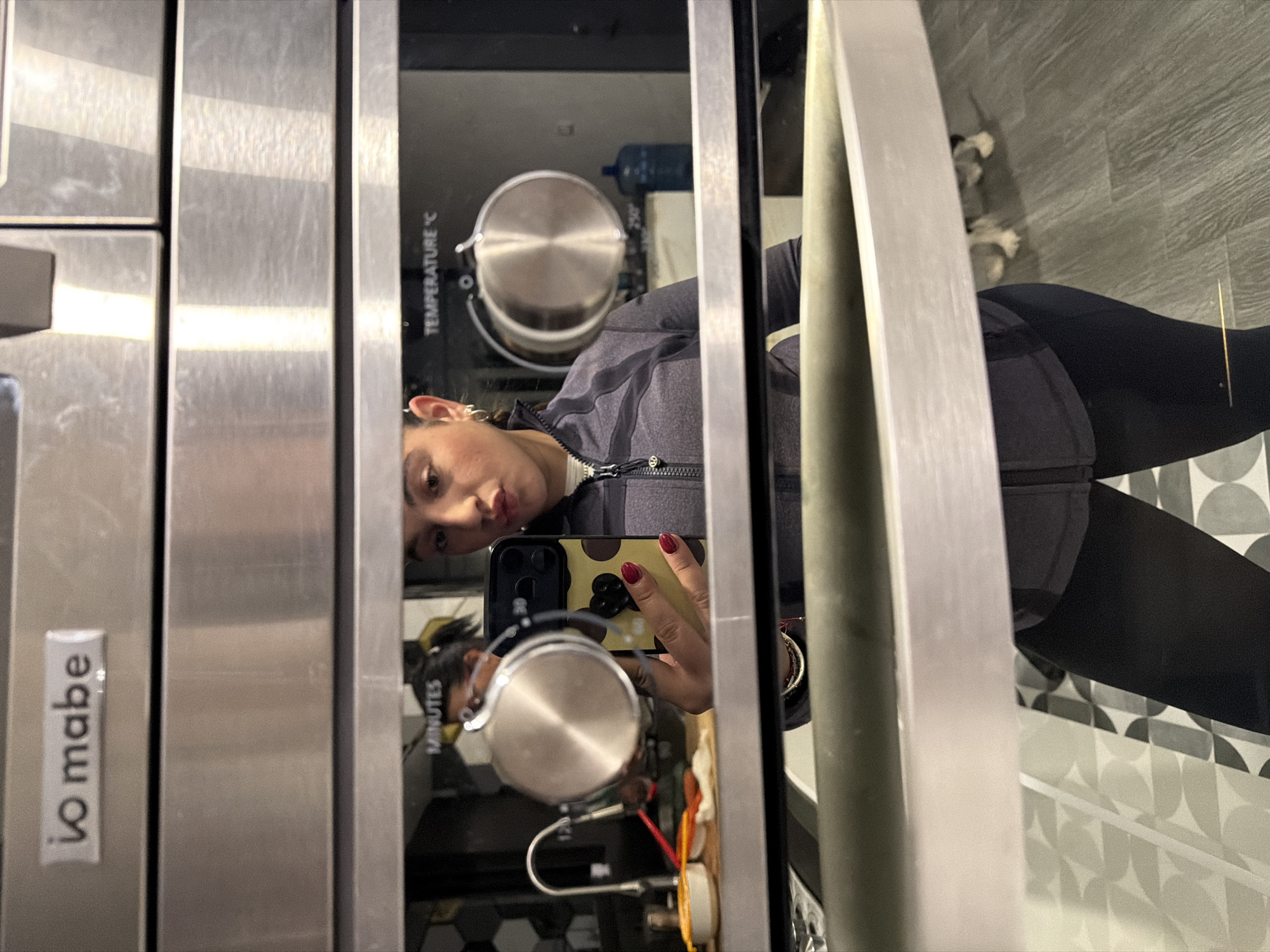 The image, originally rotated 90 degrees clockwise, displays a woman's reflection in the glass door of a modern kitchen appliance, likely an oven or microwave. The woman, appearing to take a selfie, is visible from the chest up, wearing a dark grey zip-up athletic jacket and black leggings. Her dark hair is pulled back, and she holds a smartphone with a distinct yellow and black patterned case and a black grip accessory. Her fingernails are painted dark red.
Inside the appliance's reflective glass, a stainless steel pot or bowl is visible, alongside a large blue water dispenser bottle in the background. The appliance itself is made of silver stainless steel. On its left side, the white logo "io mabe" is vertically displayed. Within the glass, the operational texts "TEMPERATURE °C" and "MINUTES" are also visible.
In the reflected background, behind the woman's legs, there's a section of patterned floor or wall tiles featuring a geometric design of light and dark grey semi-circles. Partially visible on this patterned surface is a small dog, with patches of white and black fur, seen with its front paws and part of its body reflected. The setting appears to be an indoor kitchen or utility area, consistent with the "io mabe" brand which is prominent in Mexico, aligning with the context of Puebla, Mexico.