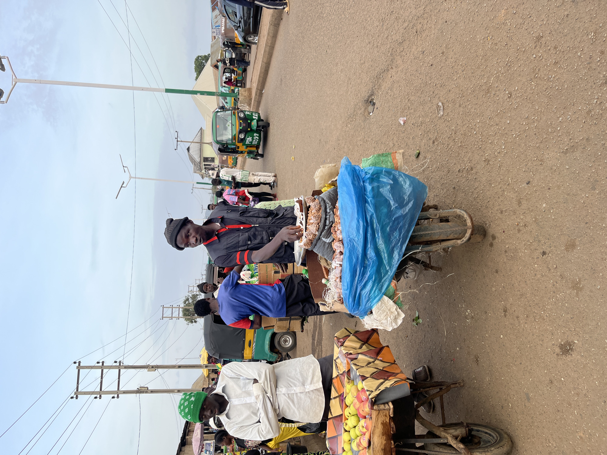 In Bukuru, Nigeria, an outdoor market scene unfolds under an overcast sky. The foreground is dominated by two vendor carts positioned on a dusty, paved road. To the left, a cart displays a selection of fresh fruit, including apples and peaches, arranged on a colorful, patterned cloth. A young man wearing a white shirt and a green beanie stands with his arms crossed beside this cart, facing slightly away from the camera.
To the right, a second vendor cart is laden with goods, partially covered by a blue plastic sheet. A man in a black vest over a black and red shirt and a black beanie stands behind this cart, holding a scoop or tool, seemingly attending to the merchandise. Behind the vendors, several people mill about, some obscured by the carts and others in the middle ground. Various vehicles, including a green auto-rickshaw (Keke Napep) with license plate PTA-11397 and other similar vehicles, are visible on the road, suggesting active local transportation. Structures, possibly market stalls or buildings, are present in the background, along with utility poles and overhead wires. The overall atmosphere depicts a bustling commercial street in a Nigerian city.