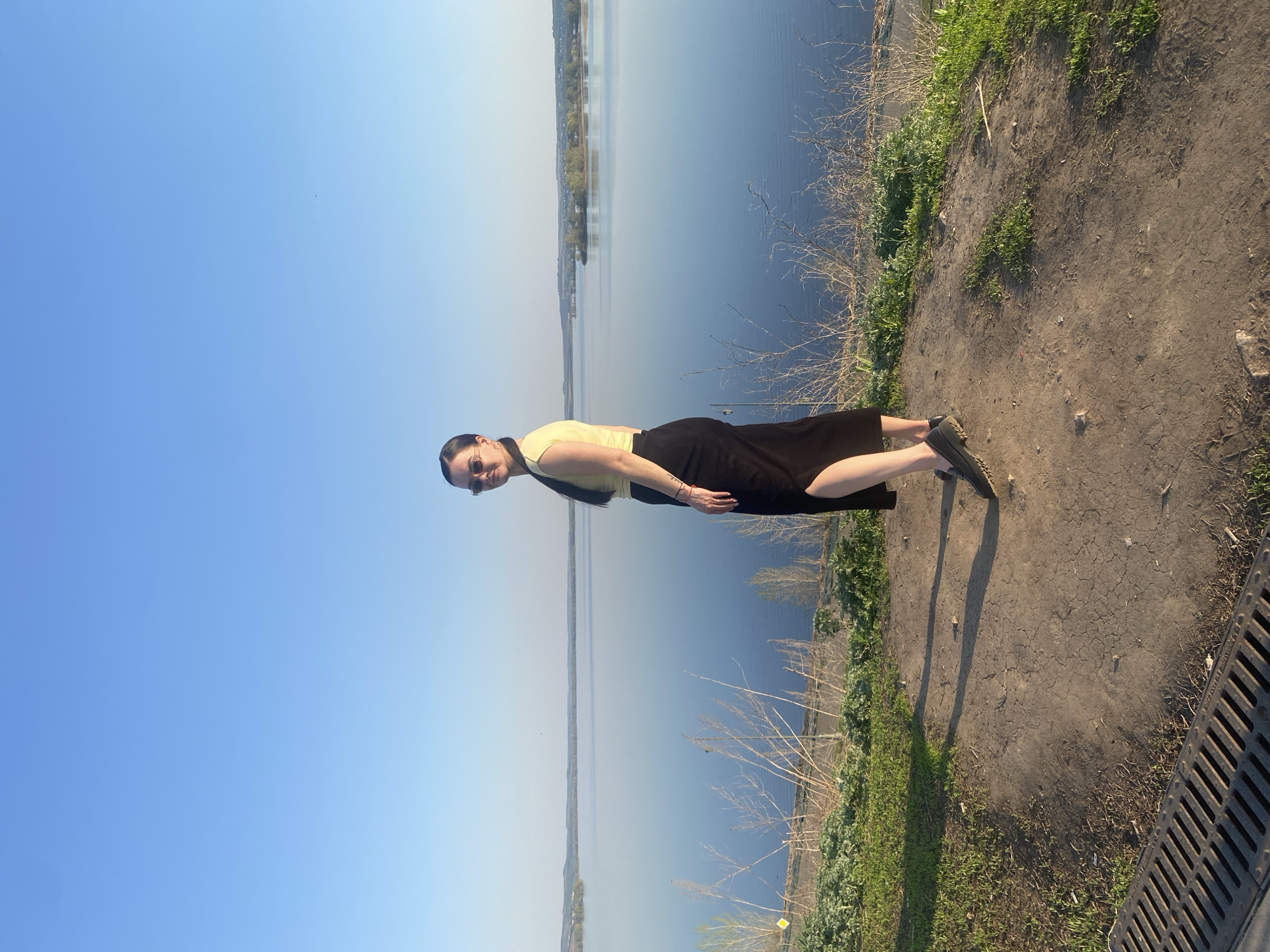 A woman is standing on the bank of the Volga River in Sviyazhsk, Russia. She has dark hair, sunglasses, and is wearing a yellow top and black skirt. The river is calm, and the sky is blue. The riverbank is grassy and has some bare branches. There is a storm drain in the lower right corner.