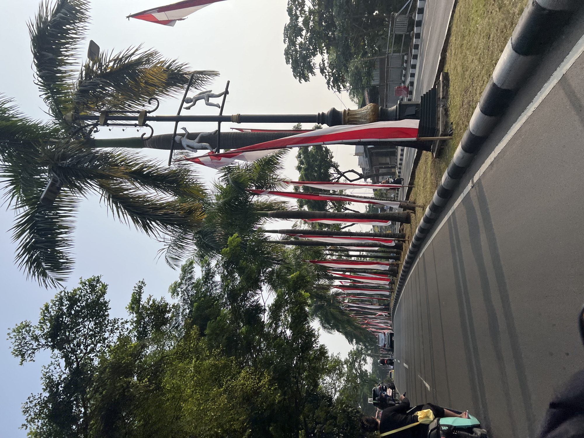 The image depicts a street scene in Cibinong, Indonesia, likely during the day given the bright lighting and clear sky. The viewpoint is from a moving vehicle, possibly a motorcycle, looking forward along a road lined with palm trees and tall, red and white flags. These flags, characteristic of the Indonesian national flag, are hung at intervals along the roadside, suggesting a patriotic or festive occasion.
In the foreground, the rear of a turquoise motorcycle is visible, with its rider wearing a black top and a yellow bag slung over their shoulder. The road is paved asphalt with white lane markings and a black and grey striped curb separating it from a grassy median. Several cars and motorcycles are visible in the distance, navigating the road. The palm trees, some of which are quite tall, line the right side of the road, and their fronds cast long shadows across the asphalt. A lamppost with two decorative sculptures of figures is also prominent. Beyond the road, there are trees and some buildings in the background. The overall impression is a well-maintained urban street decorated for an event.
