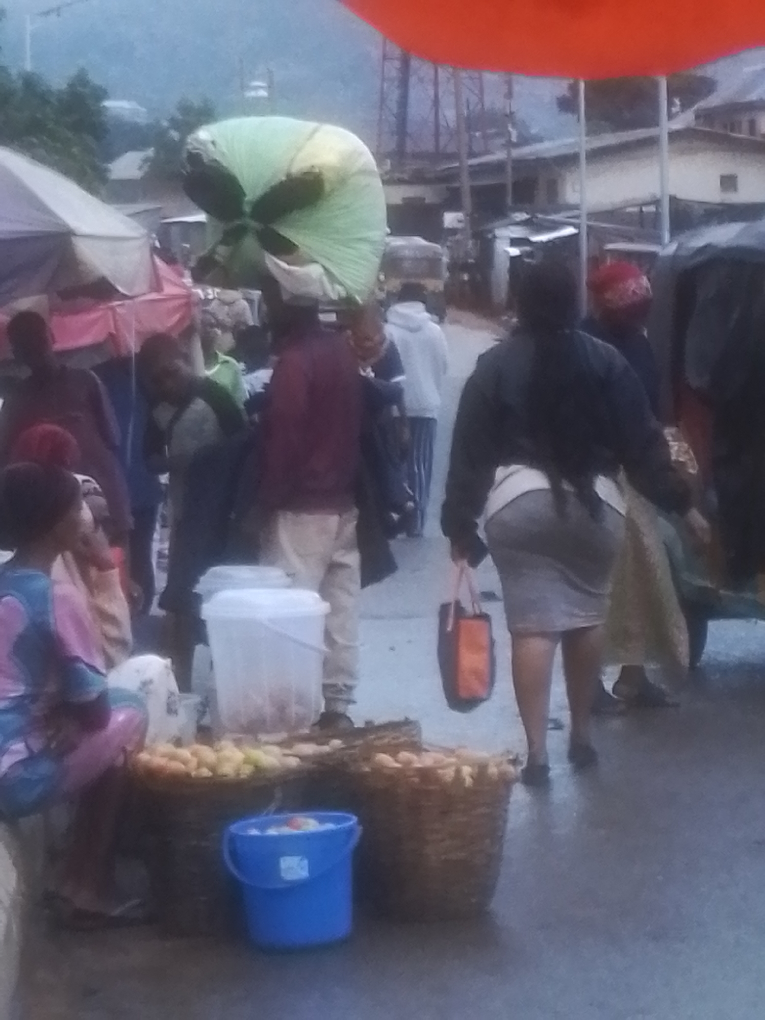 The image depicts a street market scene in Gwong, Nigeria. A person is carrying a large, light green bundle on their head. In the foreground, several baskets filled with fruits, possibly yams or plantains, are visible. A blue bucket sits next to the baskets. Other individuals are present in the background, some appearing to be vendors or customers. Tents or market stalls are set up, and buildings can be seen in the distance. The overall atmosphere suggests a busy marketplace, potentially during a cloudy or overcast day.