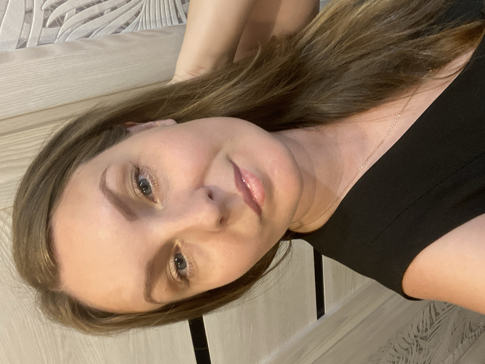 This is a close-up selfie of a woman with long, brown hair and blue eyes. She is wearing a black top and a delicate gold necklace. Her left arm is raised and behind her head, with her hand resting on her hair. She has light makeup on, including mascara, lip gloss, and blush. The background appears to be indoors, with a textured wall and a vertical striped element visible. There are no indications of the location being in Noviy, Russia.