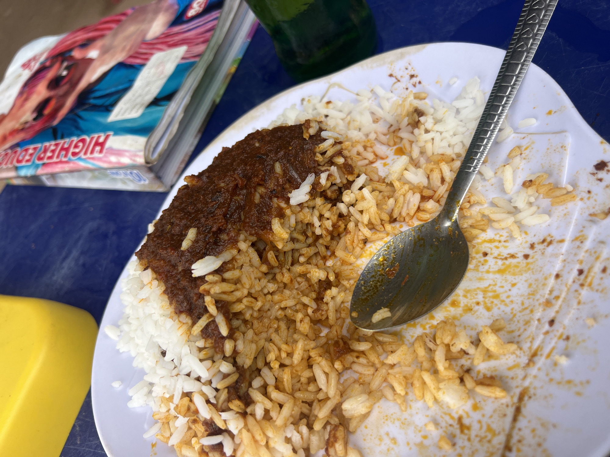 The image shows a partially eaten meal of rice and a dark stew on a white plate. A spoon rests on the plate. Part of a magazine with the text "HIGHER EDU" is visible in the upper left corner, along with a green bottle and a yellow container. The meal is on a blue surface. This image was likely taken in Jalingo, Nigeria, based on common food and cultural contexts.