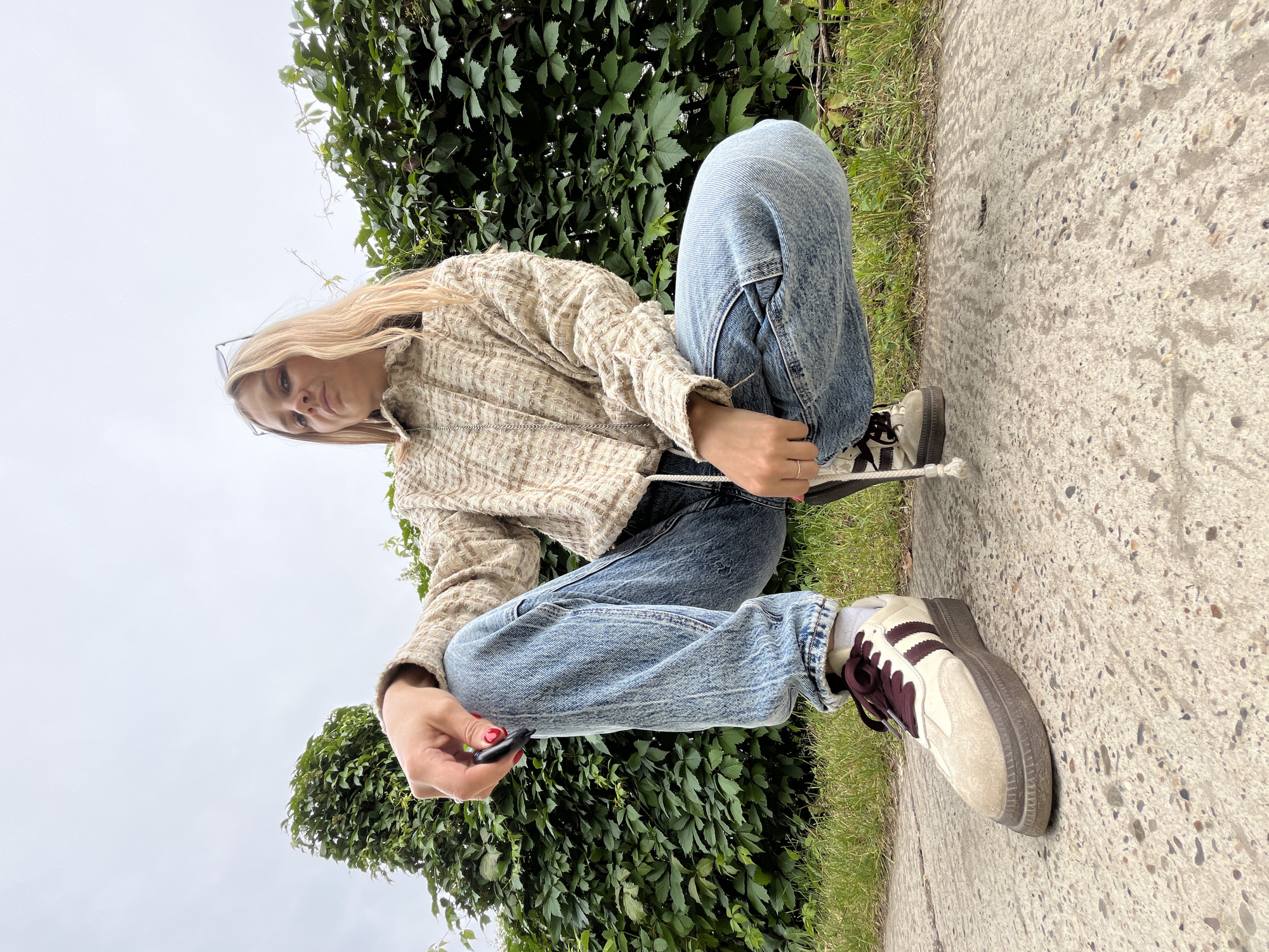A young woman with blonde hair is crouching on a concrete path next to a green hedge. She is wearing a textured, cream-colored jacket, light-wash denim jeans, and beige and burgundy athletic shoes. The woman is holding a dark object, possibly a small electronic device, in her right hand. Her left hand is resting on her knee. She is looking towards the camera with a neutral expression. Her glasses are pushed up onto her head. The background consists of lush green foliage and a pale, overcast sky, suggesting it might be a cloudy day. The concrete path has a speckled texture. The overall scene appears to be outdoors, possibly in a park or garden area.