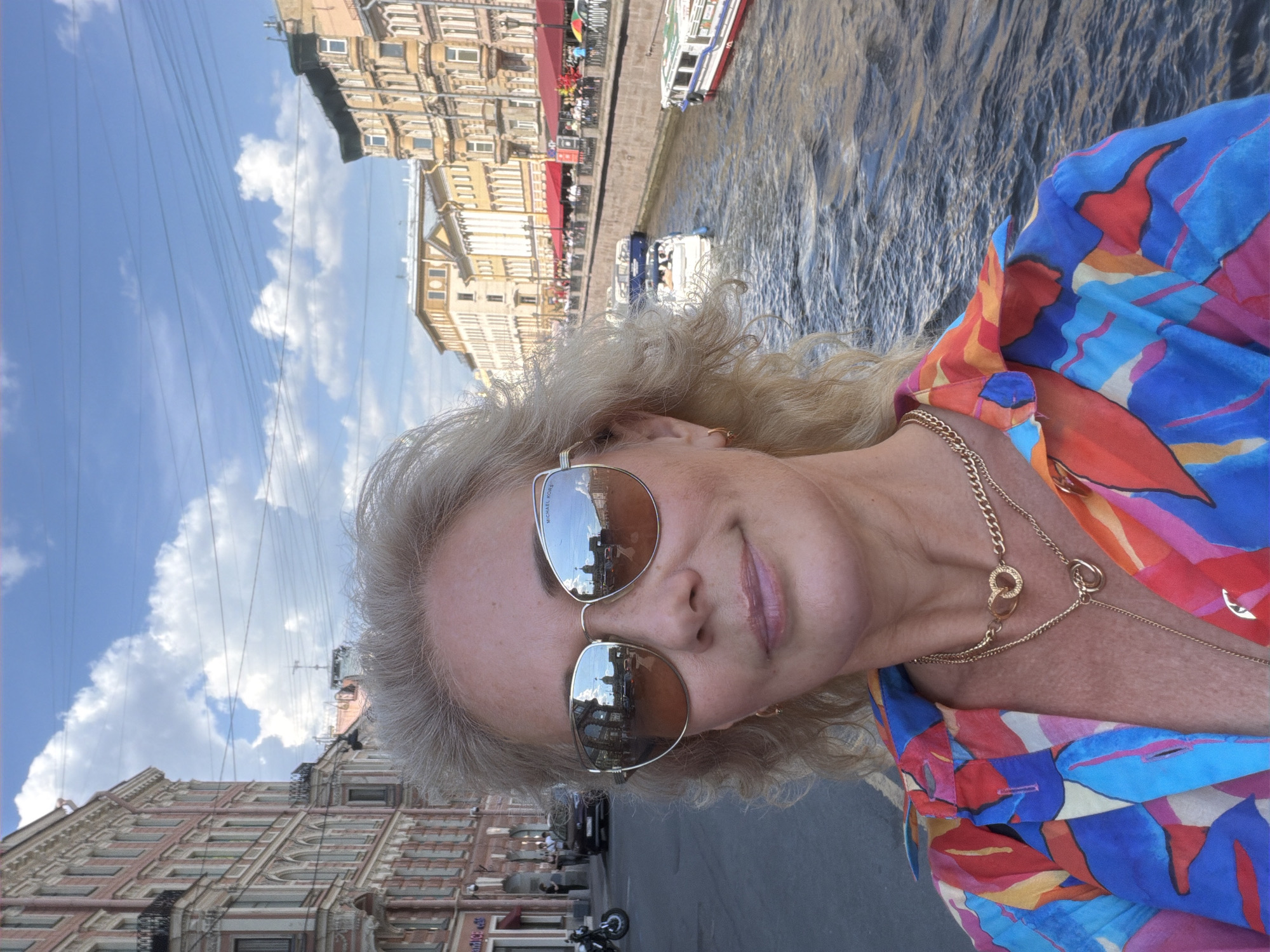 A brightly lit, daytime selfie-style portrait features a woman with light blonde hair, wearing large mirrored sunglasses and a vibrant, multi-colored patterned shirt. She has a slight smile and is adorned with multiple gold chain necklaces. The sunglasses faintly display "MICHAEL KORS" on the right lens and reflect a cityscape.
The background reveals an urban landscape along a canal or river, characteristic of Saint Petersburg, Russia. On the right, the water is dotted with several tourist boats. The banks are lined with elegant, historical buildings, some featuring red awnings or umbrellas over outdoor cafes. On the left side of the image, more buildings flank a street where a motorcycle is visible. The sky above is a clear blue with scattered white clouds, and a network of overhead power lines traverses the frame. The overall atmosphere appears sunny and pleasant.