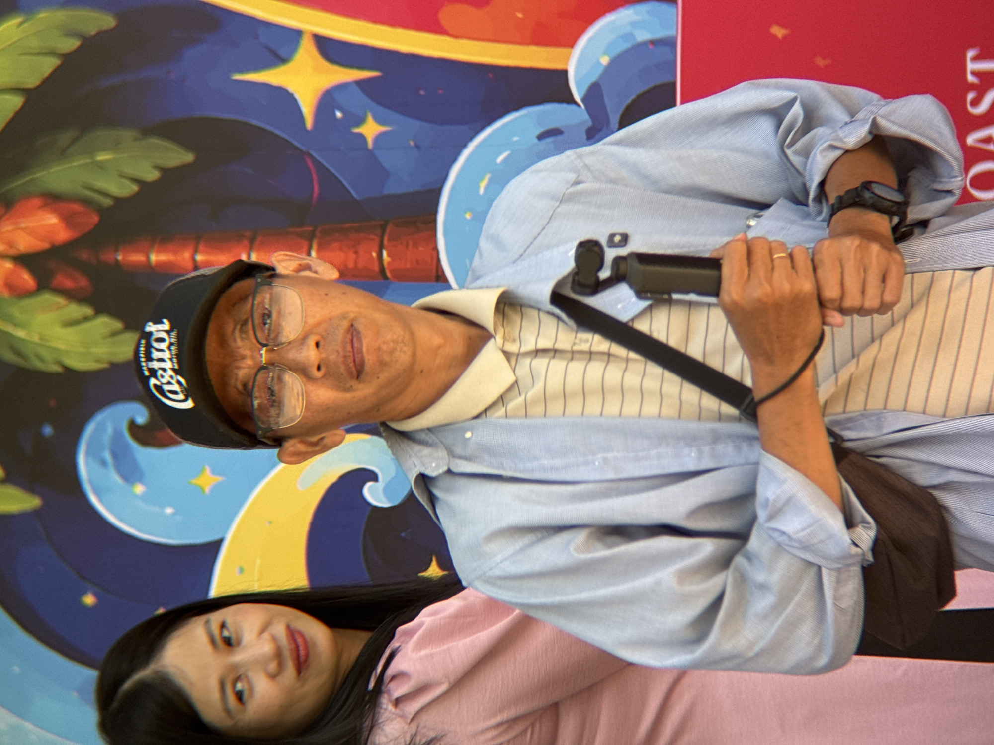 Two people are posing for a photo in front of a colorful mural. The man in the foreground is wearing a light blue button-down shirt over a striped t-shirt and a black baseball cap. He is holding a black stabilizer or gimbal in his hands. He is also wearing glasses and a gold ring on his left ring finger. A dark brown bag is slung across his chest. The woman in the background is wearing a pink top and has long black hair. She is smiling at the camera. The mural behind them features a tropical theme with palm trees, stylized waves, and stars on a dark blue background. There is text visible on the red board behind the man, but it is mostly obscured. The image appears to be taken during the day. The overall atmosphere seems cheerful and relaxed, suggesting a tourist or recreational setting in Nha Trang, Vietnam.