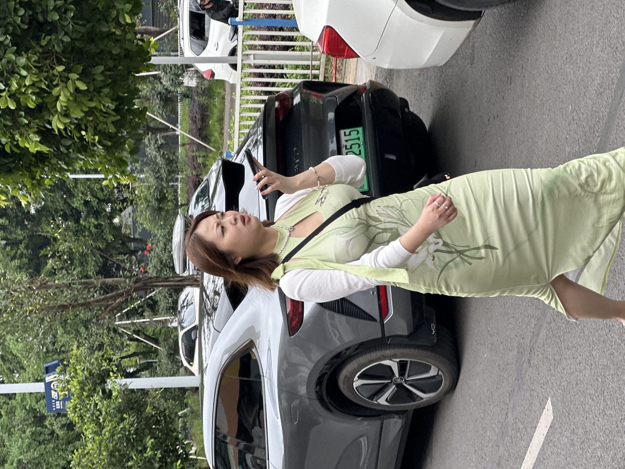 In Nan'an District, China, a woman stands on a paved surface next to parked vehicles. She is holding a mobile phone. Her dress is light green, featuring a floral design. She is wearing a white top underneath. The background includes trees and other cars. One of the vehicles nearby has a green license plate with the number 2515 visible.