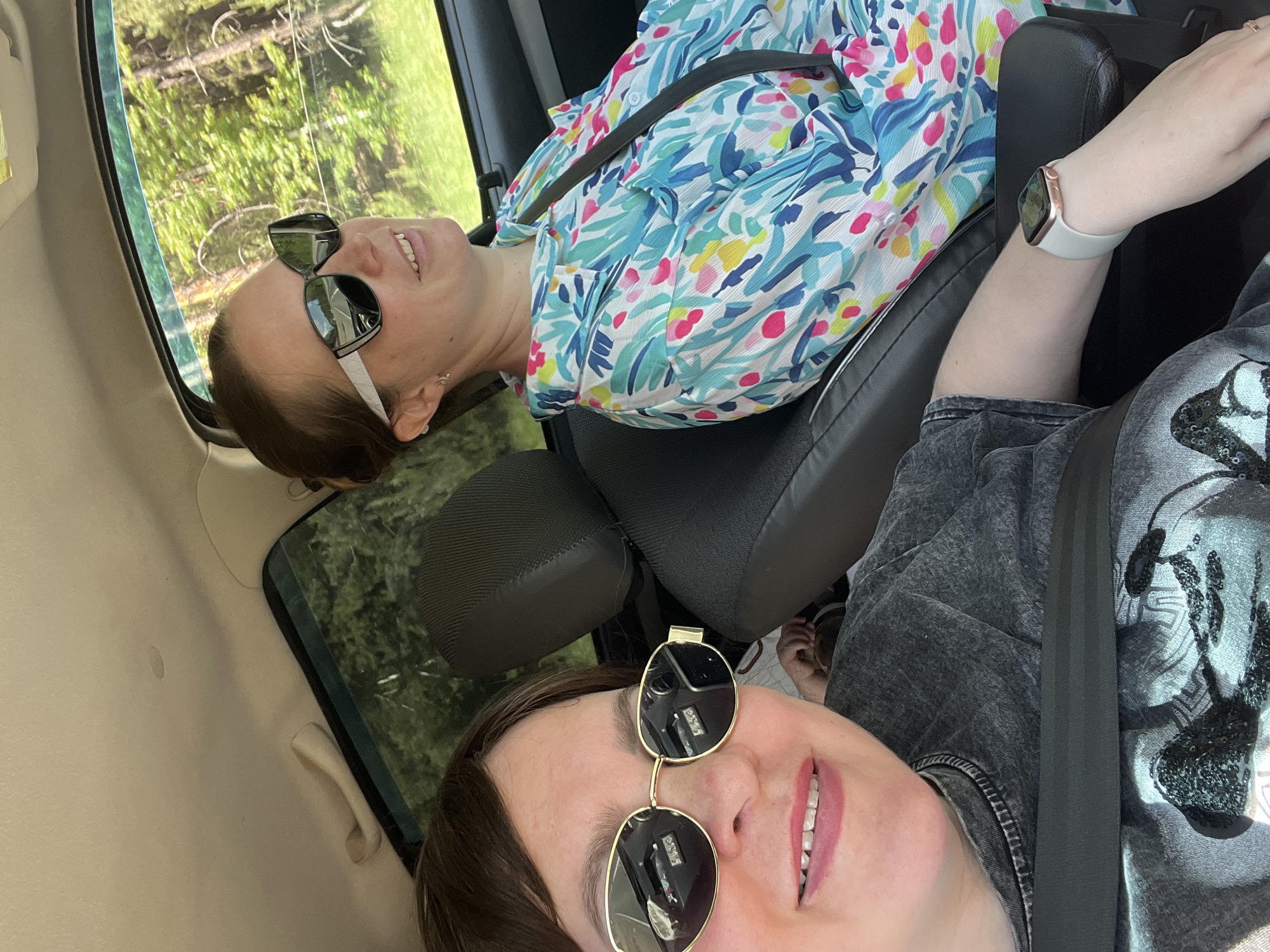 Two women are in a car. The woman in the foreground is wearing a grey shirt and gold-framed sunglasses. The woman in the background is wearing a brightly colored shirt and black sunglasses. Both women are wearing seatbelts. The location is likely rural based on the trees visible through the car window. The specific city in Russia cannot be determined from this image.