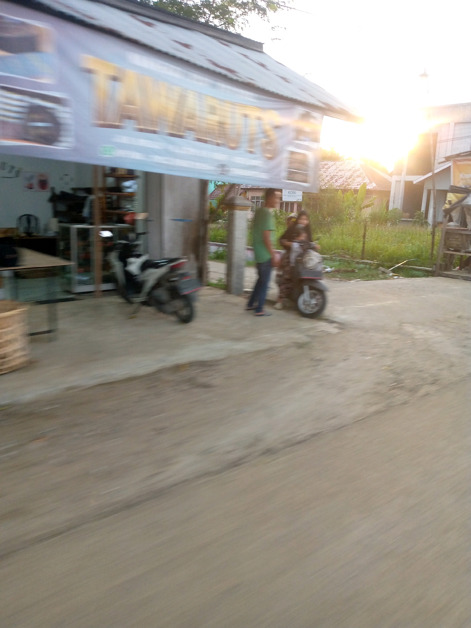 In Keudeteupinpunti, Indonesia, an outdoor scene shows a blurred view of a building with a metal roof and a large banner displaying text. Two individuals are present. One, wearing a green shirt and dark pants, stands to the left. To the right, another person is seated on a scooter. A parked motorcycle is visible in front of the building. The ground is paved, and vegetation lines the background. The sun is bright, casting a strong glare on the right side of the image.