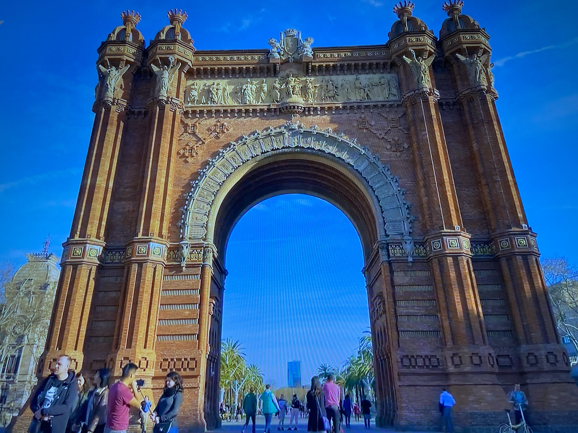 The image shows the Arc de Triomf in Barcelona, Spain. It is a large, brick structure with an archway in the center. People are walking around the arch. A building is visible in the distance through the archway. The sky is blue.