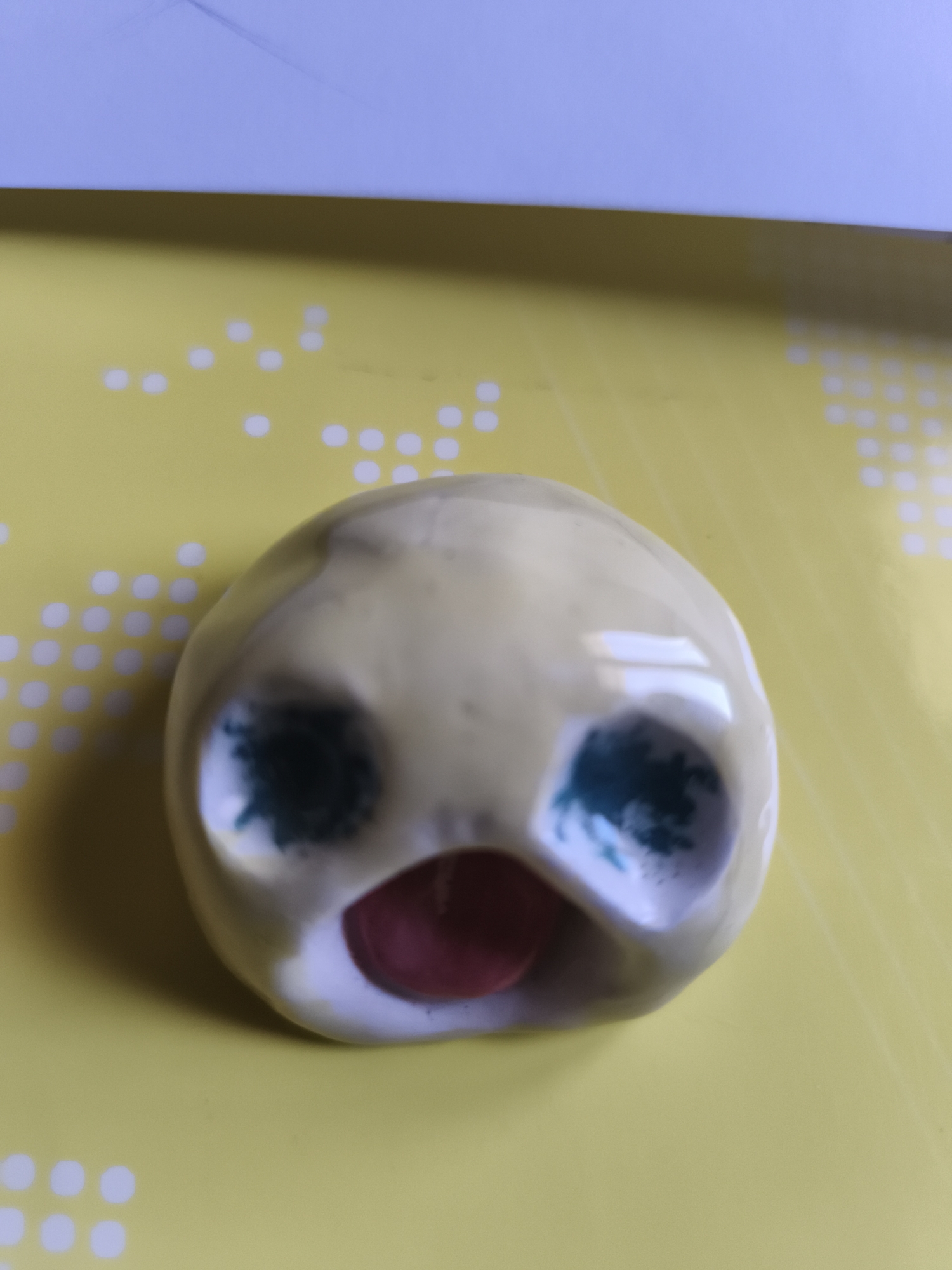 A close-up, high-angle, slightly blurred shot shows a yellow, oval-shaped ceramic object with a gaping mouth and two large, round, dark green eyes. The object is resting on a bright yellow surface with a faint white dotted pattern. A light blue surface is visible at the top of the frame. The ceramic object appears to be a stylized face, possibly a caricature or a decorative item. There is no indication of location in Saint-Denis, France.