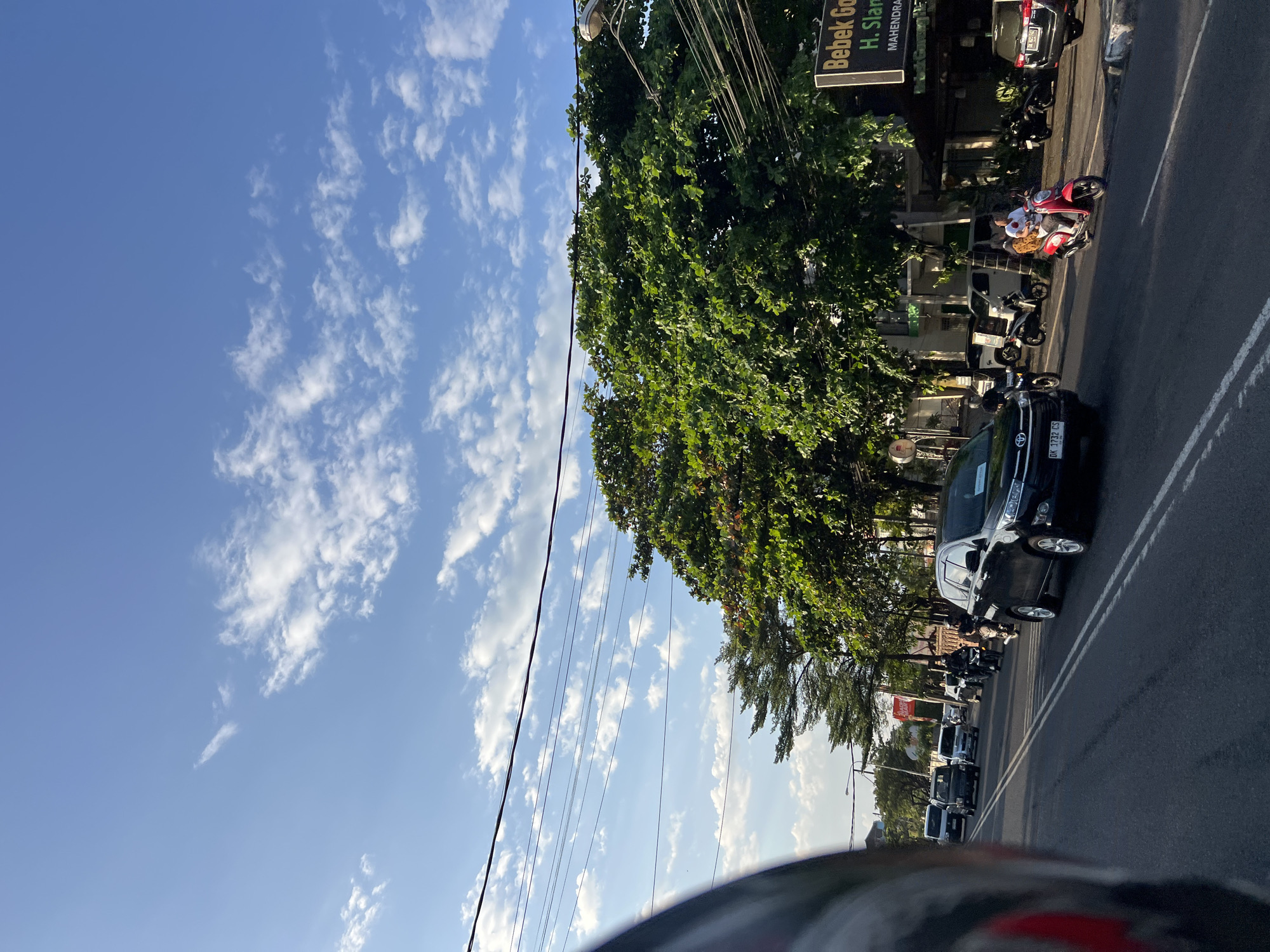 The image captures a street scene in Denpasar, Indonesia, under a bright blue sky with scattered clouds. The perspective is from a moving vehicle, with the left side of the frame partially obscured by the edge of a helmet.
In the foreground, the asphalt road displays double white lines, indicating multiple lanes of traffic. Several vehicles are visible on the road, including a black SUV in the right lane, followed by a red scooter carrying two people, and further down, a line of other cars and motorcycles. The vehicles appear to be moving in the same direction.
On the right side of the road, a commercial building with a sign reading "Bebek Goreng H. Slamet" and "MAHENDRAN" is partially visible. This building is shaded by lush green trees with dense foliage. Electrical wires are strung overhead, crossing the sky.
On the left side of the road, more vehicles are seen, including a white van and other cars. Another set of tall, green trees lines the left side of the street, casting shadows on the road. The general environment suggests a busy urban street in Denpasar. The lighting indicates it is daytime, likely late afternoon given the angle of the sun and the warm tones on the clouds.