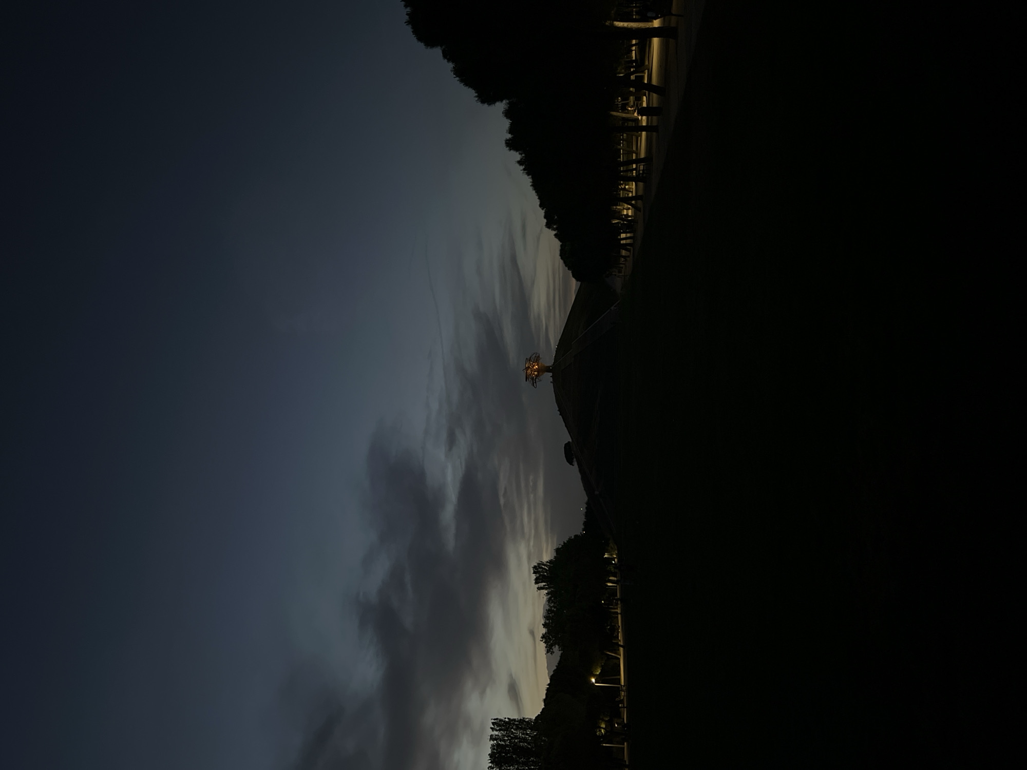 At dusk, the image captures a scene in Madrid, Spain, showing the silhouette of the Temple of Debod against a darkening sky with scattered clouds. The temple is illuminated by warm lights. A park is visible on the right side of the image, with trees lining the edge and benches sparsely arranged along a walkway. The left side of the image is dominated by the fading light of the sky.