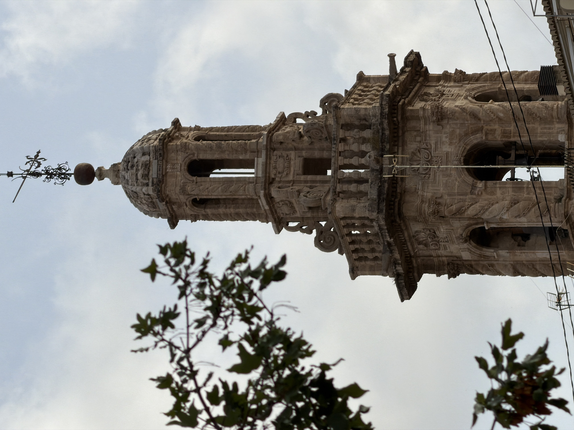 An ornate, weathered stone bell tower with carved details stands against a cloudy sky. The tower has multiple levels, with arched openings and decorative stonework. At its apex is a weather vane. To the left, a branch of a leafy tree, slightly out of focus, hangs in the foreground. Electrical wires are visible crossing the lower portion of the tower. The overall impression is of an old, historic structure in an urban setting.
