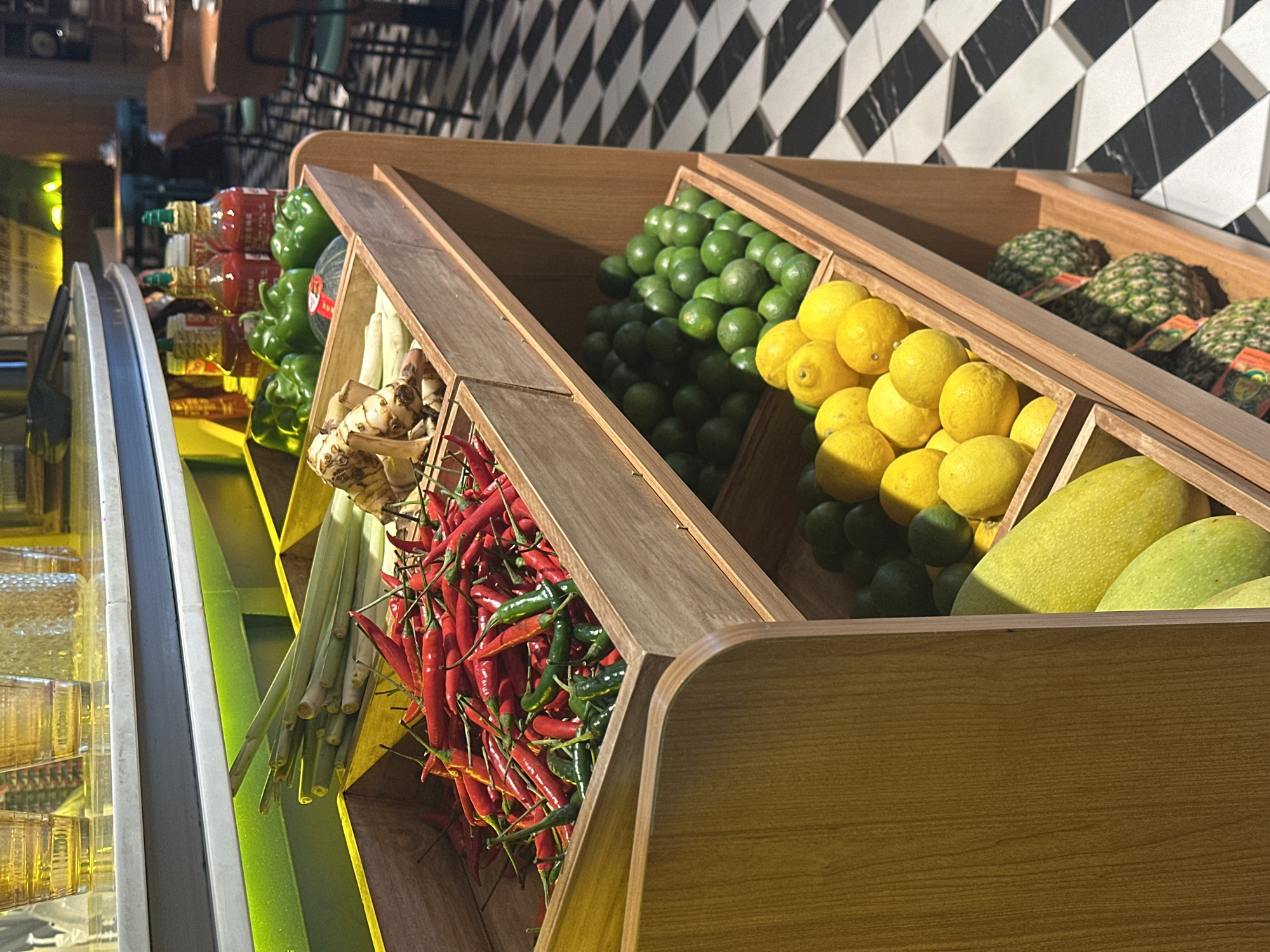 This image shows a wooden display shelf with various fruits and vegetables, located in a restaurant in Futian District, China. The top shelf contains lemongrass and galangal. The second shelf holds a pile of red chili peppers and some green ones, as well as green bell peppers and what appears to be a small pumpkin or squash. The third shelf displays a collection of limes and lemons. The bottom shelf contains two large mangoes, and below them are three pineapples. In the background, the restaurant setting is visible, with tables and chairs, and a black and white patterned floor.