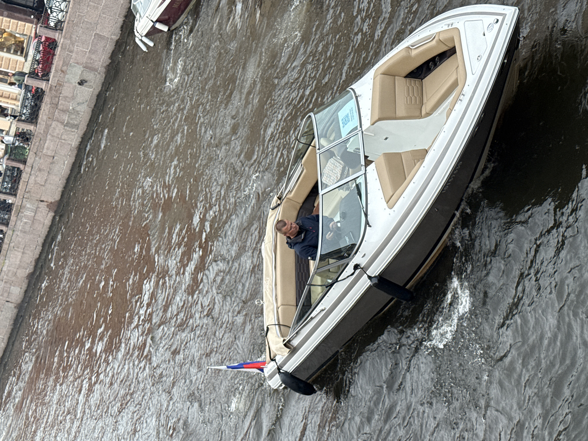 A man is operating a modern white and beige motorboat on a canal in Saint Petersburg, Russia. The boat is sleek with upholstered seating. A Russian flag is attached to the stern. The man appears to be looking ahead, focused on navigating the boat. The water is a murky brown and appears choppy, suggesting movement. Along the left side of the canal, a stone embankment with decorative railings is visible, hinting at an urban setting. The overall atmosphere seems to be one of leisure or transport on the water. The time of day appears to be daytime, and the weather is overcast or hazy, as there are no strong shadows.