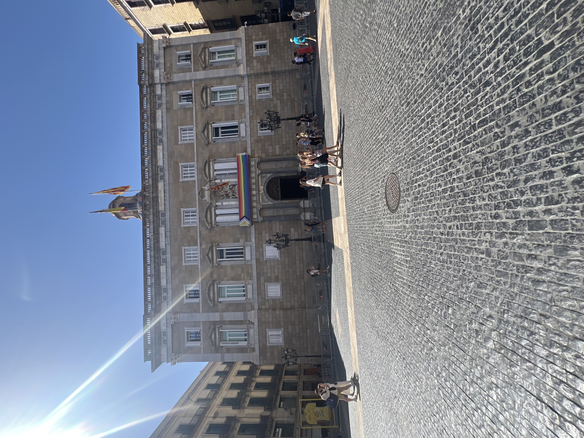 The image depicts a sunny day in Barcelona, Spain, specifically in front of the Palau de la Generalitat de Catalunya on Plaça Sant Jaume. The building is a grand, historical structure with a light-colored stone facade, adorned with multiple windows and a decorative cornice. Two flags, one with yellow and red stripes and another with yellow and red vertical bands, fly from the roofline. A rainbow banner is displayed prominently across the building's central section.
The foreground is dominated by a cobblestone plaza, with a distinctive pattern of grey stones. Several individuals are present in the scene. In the middle ground, a group of people walks across the plaza. To the left, two people are standing and interacting. To the right, more individuals are walking, some in casual attire. Street lamps and metal barriers are also visible within the plaza. The sky is clear and blue, with bright sunlight casting strong shadows. The overall atmosphere suggests a public gathering or a typical day in a busy city square.