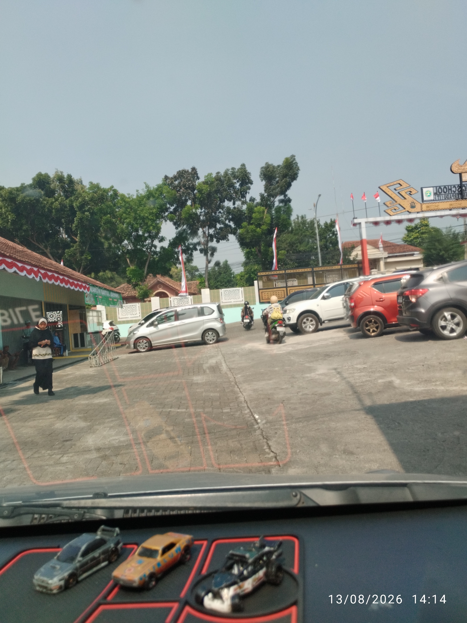The image shows an outdoor scene of a parking lot in front of a building with Indonesian flags displayed. In the foreground, a car's dashboard is visible, with three toy cars placed on a mat. The background features several parked cars, a few motorcycles, and a building with red and white decorative trim. A woman in a hijab is walking in the parking lot, and several other people are visible near the building's entrance. Trees line the area behind the building. The timestamp "13/08/2026 14:14" is overlaid on the bottom right of the image. The environment appears to be a sunny day in a suburban or semi-urban area, possibly near a school or public facility, given the flags.