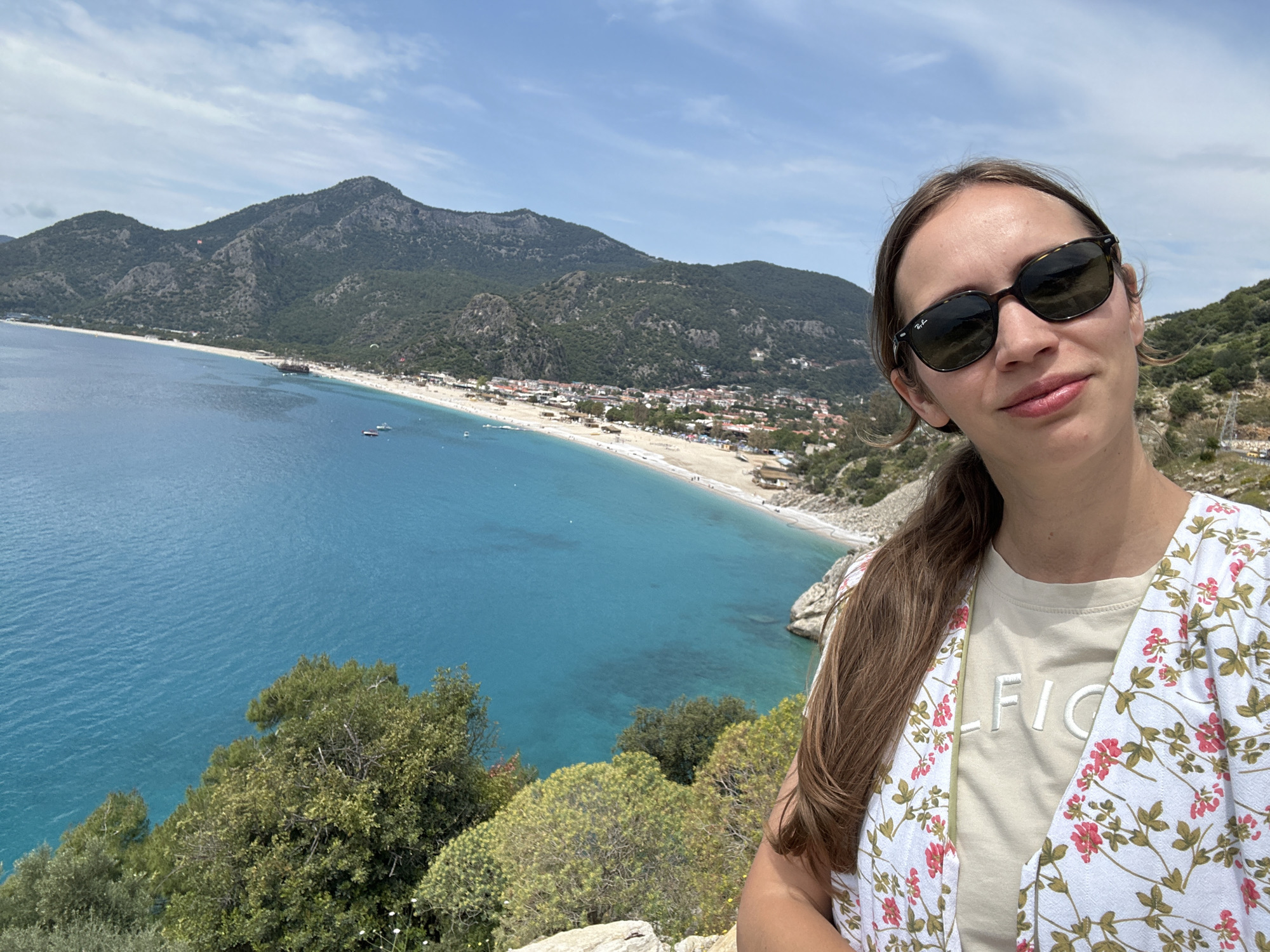 A woman is taking a selfie at Ölüdeniz, in Fethiye, Turkey. She is wearing sunglasses and a floral blouse over a tan T-shirt. Behind her is a picturesque view of the turquoise sea, a sandy beach, and a town nestled at the foot of a mountain. Some boats are visible on the water. Green vegetation is in the foreground near the woman. The sky is blue with some clouds.