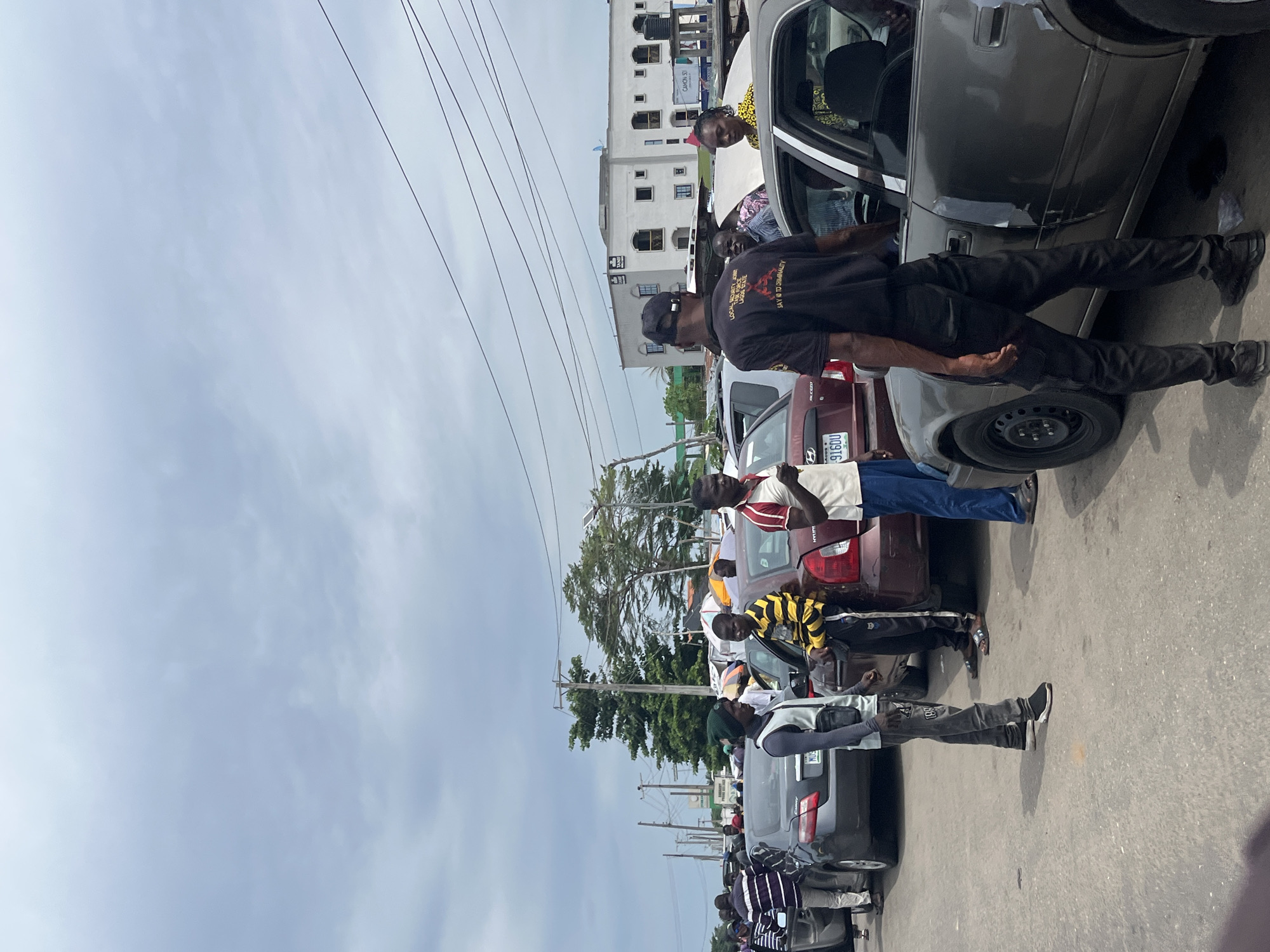 The image depicts a street scene in Badagry, Nigeria. A line of parked cars is visible along the side of the road. A group of men are standing near the vehicles. Some of the men appear to be interacting with the occupants of the cars. The sky is overcast. In the background, a multi-story building is present.