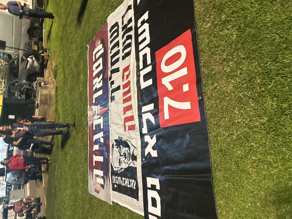 The image depicts an outdoor nighttime scene in Mishmar Hasharon, Israel, on a grassy public area illuminated by artificial light. Three large fabric banners are laid horizontally on the grass in the foreground. The uppermost banner features a partial image of a man and Hebrew text translating to "He is treason." The middle banner displays a stylized black and white portrait of Benjamin Netanyahu, accompanied by Hebrew text that translates to "Prime Minister resign" and "Opposition." The lowermost banner is black with white Hebrew text on the left, translating to "and we will not forget and will not forgive," and a prominent red rectangle on the right containing the white numerical date "7.10."
In the mid-ground, a woman in a purple top and blue jeans stands behind the banners, actively speaking into a microphone positioned on a stand. To her right, another woman in a red top and dark pants stands further back. To her left, a man in a black shirt is partially visible. In the background, on a paved surface, multiple vehicles are parked, including a silver SUV and a white cargo van. Near the vehicles, additional individuals are observed: a person crouched beside the SUV, a man in a light blue shirt holding papers near the white van, and a person seated on what appears to be a mobility device or motorcycle on the far left. Sound equipment is also visible near the parked vehicles. The scene captures a public gathering or protest focused on political messaging related to the events of October 7th.
