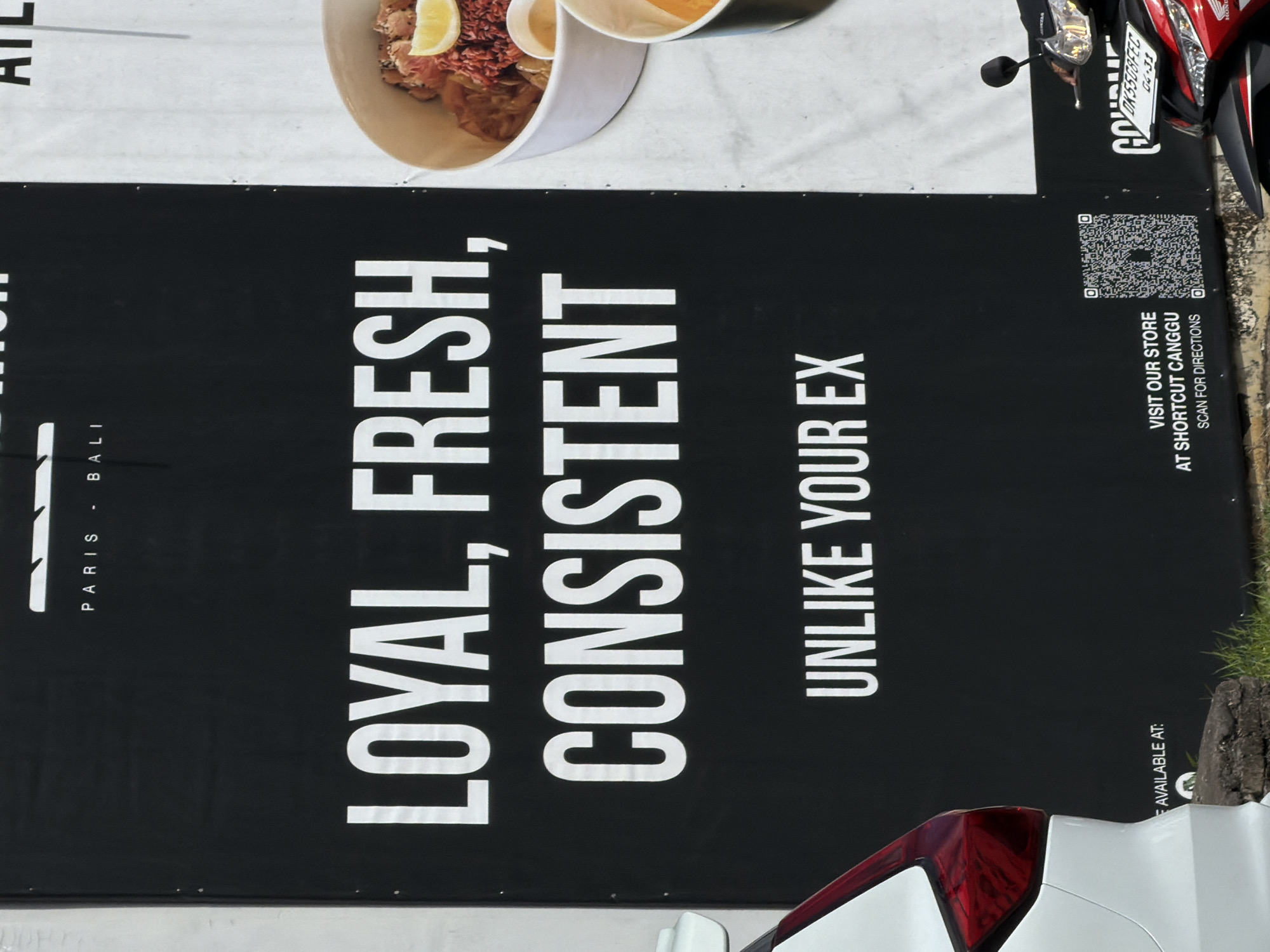 The image shows a poster or sign for a business, located in North Kuta, Indonesia. The poster reads "PARIS - BALI LOYAL, FRESH, CONSISTENT UNLIKE YOUR EX" in large, white letters against a black background. In the upper portion of the poster, there is a food item. A QR code is printed on the right side with the message "VISIT OUR STORE AT SHORTCUT CANGGU SCAN FOR DIRECTIONS". The bottom portion of the image is partially obstructed by a white vehicle. Part of a red and white scooter is visible at the top of the image, displaying the license plate number DK5508FEC. Part of a tree trunk is visible on the right side of the poster.