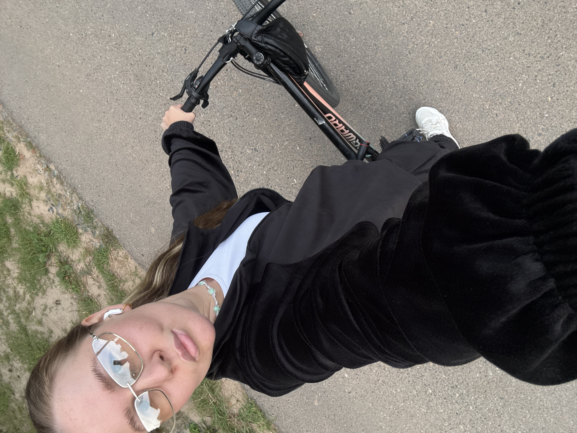 A young woman with light brown hair pulled back into a ponytail is taking a selfie while riding a bicycle. She is wearing a black velour tracksuit, a white t-shirt, and white sneakers. She has on large, rectangular, gold-rimmed sunglasses and white wireless earbuds. A light blue beaded necklace is visible around her neck. The bicycle is black with pink accents and the brand name "FORWARD" is visible on the frame. A black fanny pack is attached to the handlebars. She is holding the handlebars with her right hand. The background shows a paved path next to a grassy area. The scene appears to be outdoors in a park or on a bike path, possibly in Chaikovsky, Russia, given the context. The lighting suggests it is daytime.