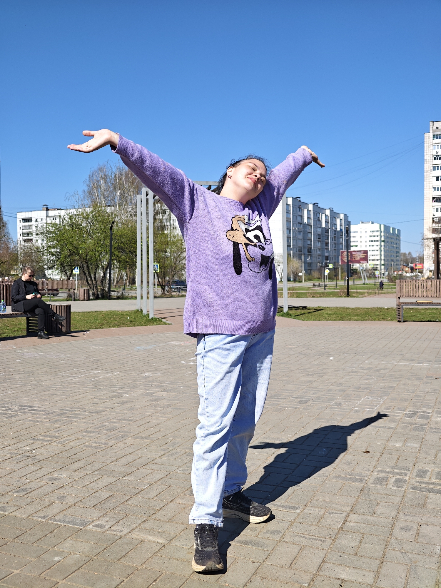 A young woman stands in a paved public space, arms outstretched, head tilted back in what appears to be a moment of enjoyment or relaxation. She wears a purple sweater with a cartoon character on it, light blue jeans, and dark sneakers. The pavement is made of gray bricks. Her shadow is cast on the ground in front of her.
In the background, there are apartment buildings and some trees. To the left, a man is sitting on a park bench, seemingly looking at his phone. Other benches are visible nearby, as well as metal structures. The sky is clear and blue.
The location is most likely in the city of Chaikovsky, Russia, as indicated by the prompt.