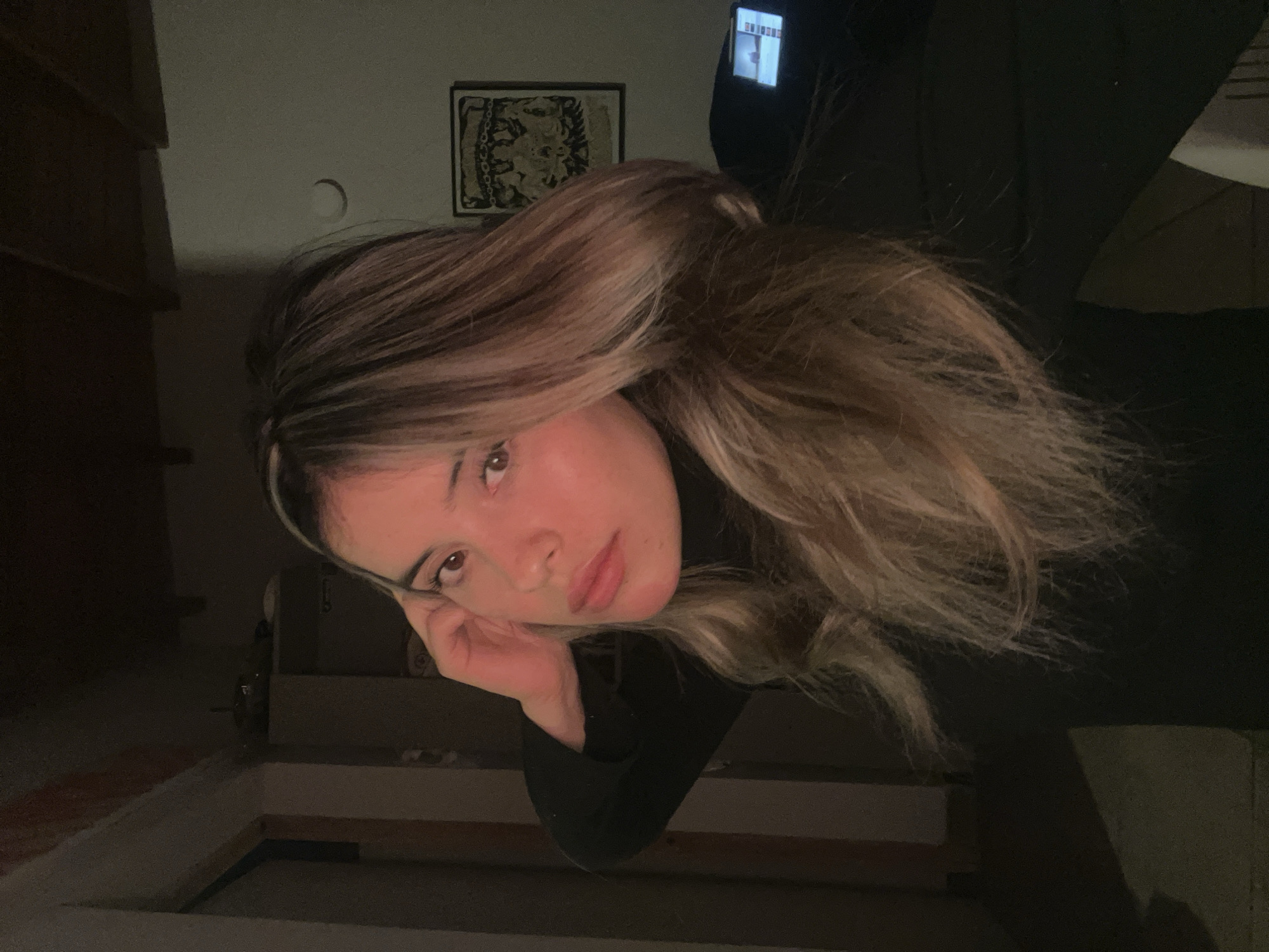 This is a low-angle, close-up selfie of a young woman in Sumalao, Argentina. She has long, layered blonde and brown hair, with bangs. Her left hand is resting on her cheek, and she is looking directly at the camera. She is wearing a black turtleneck.
The background is dimly lit, with a few objects visible. To the left, there appears to be a wooden dresser or shelf. On the wall behind her, there is a framed tapestry or artwork with intricate designs. To the upper right, a cell phone is propped up, displaying a bright screen. The overall lighting suggests it is nighttime or indoors with artificial light. The mood of the image is somewhat contemplative, with the woman's gaze and posture conveying a sense of introspection. There is no discernible text in the image.