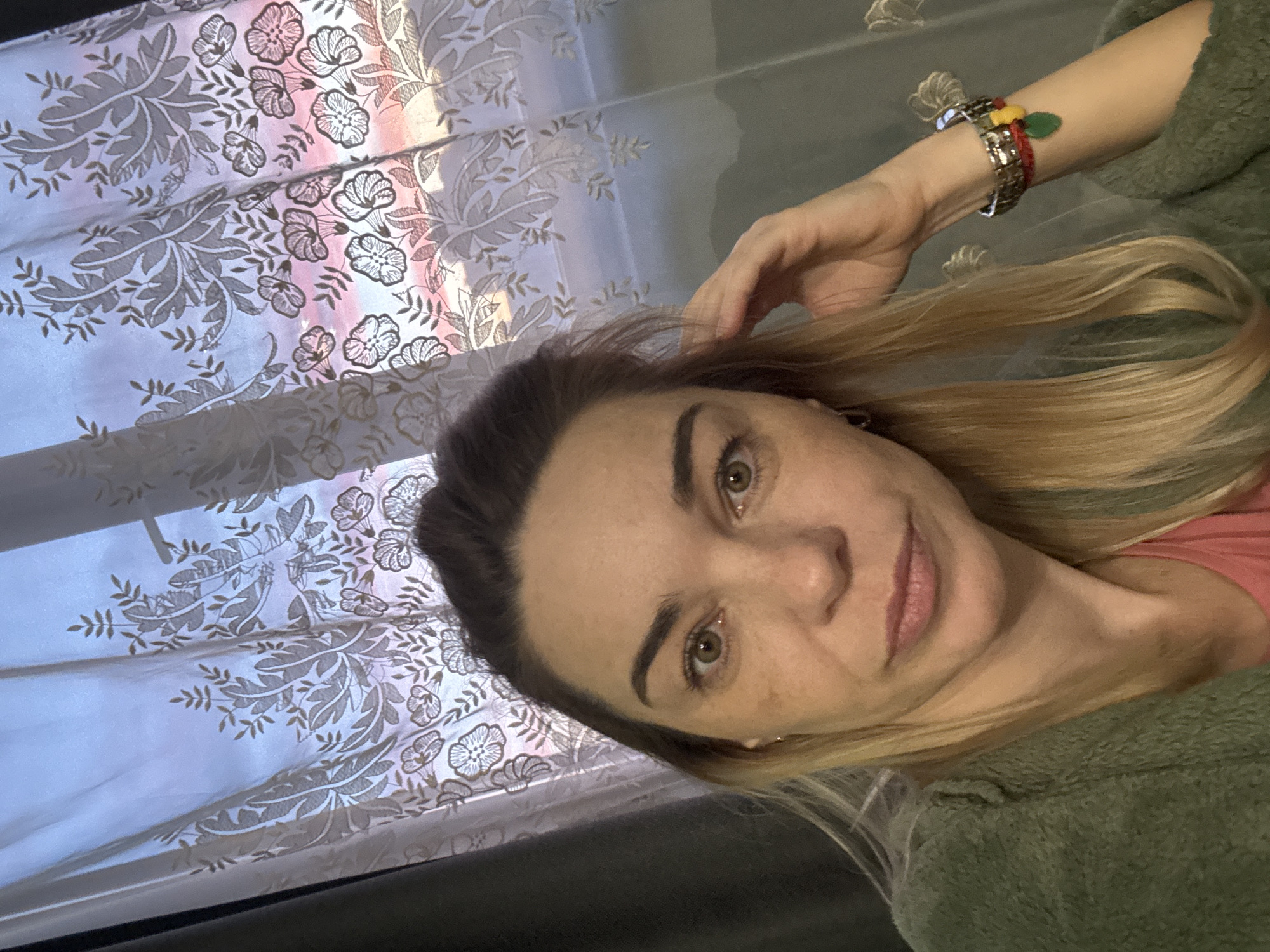 A fair-skinned woman with light brown hair and hazel eyes is seen in a selfie, positioned at a tilted angle. She wears a green garment and has bracelets on her left wrist. Her left hand is touching her hair. The background features a patterned curtain illuminated by an external light source. The image was taken indoors. There is no evidence to place the location of the image in Ladozhskoe Ozero, Russia.