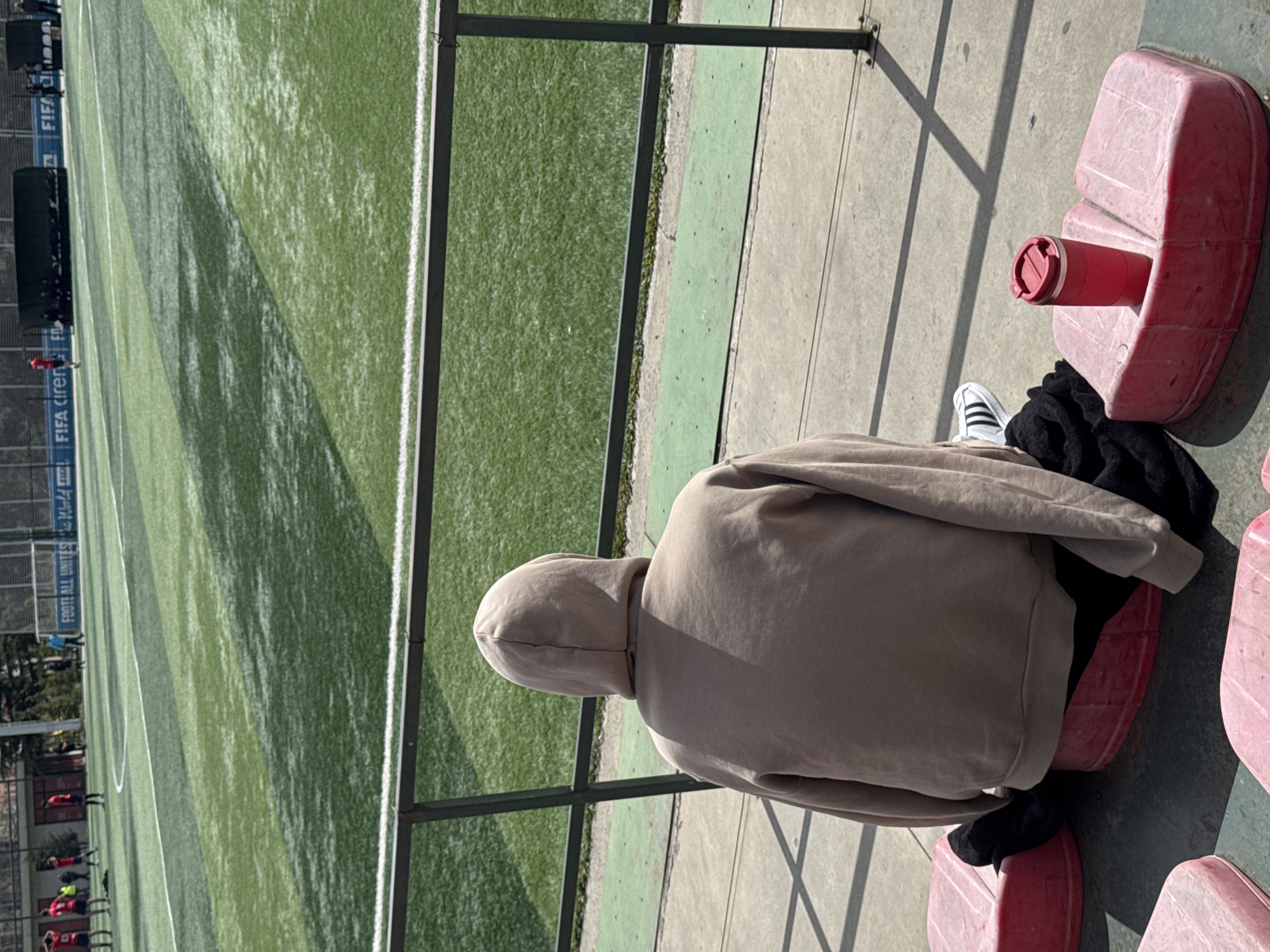 A person wearing a beige hoodie is sitting on a red plastic seat at a soccer field in Tbilisi, Georgia. The person is viewed from behind, and their head is covered by the hood. They are wearing black socks and white Adidas shoes. A red thermos is placed inside the plastic seat beside the person. A green soccer field is visible behind a fence, with players in red jerseys visible in the background. FIFA signage is seen on the fence, and the text "FOOTBALL UNITES" is visible on a sign.