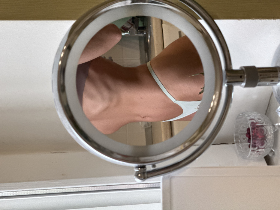 The image presents a close-up, waist-level reflection of a person in a round, chrome-framed magnifying mirror on a stand. The reflected individual, appearing female, is seen in side profile, displaying a lean physique with visible ribs and a tattoo of green foliage on the upper right thigh/hip area. They are wearing light blue G-string style underwear. The reflection reveals a background consistent with a kitchen or bathroom, featuring a sink and a metal faucet. Outside the mirror's frame, on a surface to the left, a clear, footed glass bowl contains numerous red berries. A white window frame or door jamb is partially visible on the far left. The lighting suggests an indoor setting with natural light source. No discernible visual information in the image allows for the identification of a specific location within Saint Petersburg, Russia.