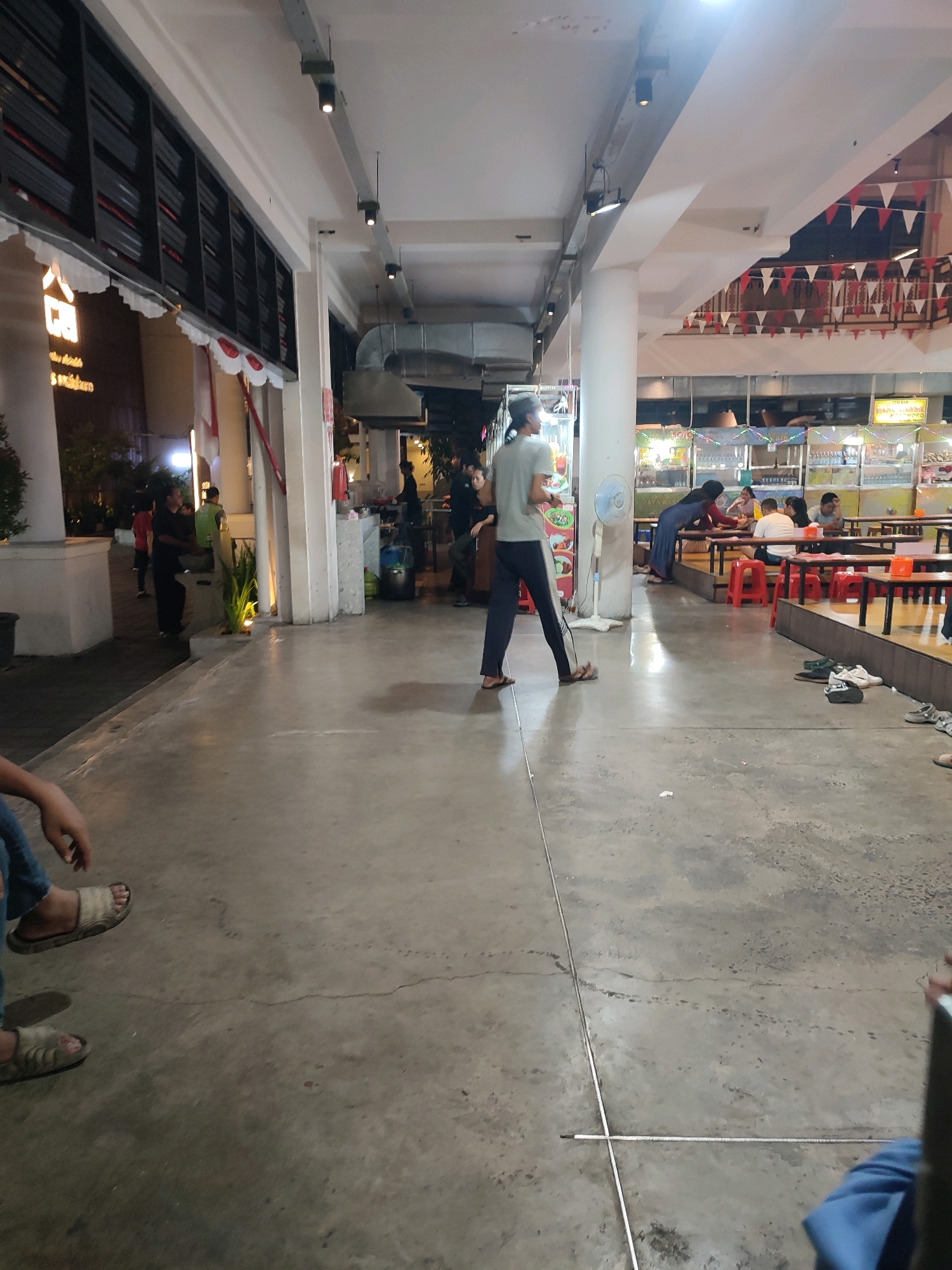 This is a medium shot taken at night of an outdoor food court, likely in Yogyakarta, Indonesia, given the signage in the background. The food court is open-air, with a concrete floor and a white ceiling supported by large white columns. Overhead lighting fixtures are visible, and a fan stands in the middle ground. Several food stalls are set up along the back wall, some with colorful lights and signage. In the foreground, a person wearing jeans and sandals is partially visible, sitting down. A person in the mid-ground is walking away from the camera, wearing a light-colored t-shirt and dark pants. Several other people are seated at tables in the background, appearing to be eating or socializing. Red plastic stools are scattered around the tables. The overall atmosphere is casual and bustling, typical of a night market or food court.