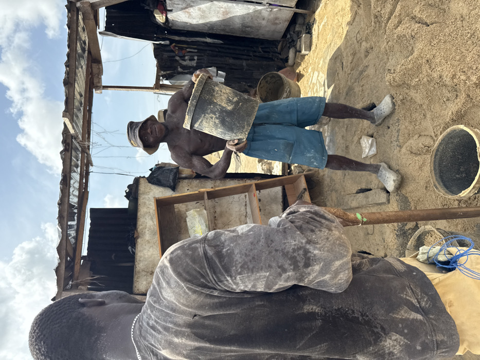The scene, set in Ado, Nigeria, depicts an outdoor work environment under a partially cloudy sky. In the foreground, a dark-skinned male with his back to the camera wears a heavily stained dark gray long-sleeved sweatshirt and light brown trousers, holding a long-handled tool vertically. A silver chain is visible around his neck. At his feet is a small blue bucket containing material and blue string. In the midground, facing the camera, a dark-skinned shirtless male wears a dark bucket hat, light blue shorts, and light-colored slip-on shoes. He holds a dirty black plastic bucket with both hands, presenting it forward, and displays a slight smile. His skin and clothing are visibly covered in light-colored residue. The ground is composed of uneven, light-colored sand or fine dirt, with an empty black plastic bucket near the foreground figure's feet and another behind the midground figure. The background features dilapidated, makeshift structures constructed from corrugated metal sheets, wood, and possibly concrete blocks, with exposed wooden beams forming an overhead frame. To the left of the midground figure, a simple wooden shelving unit with multiple empty shelves is visible.