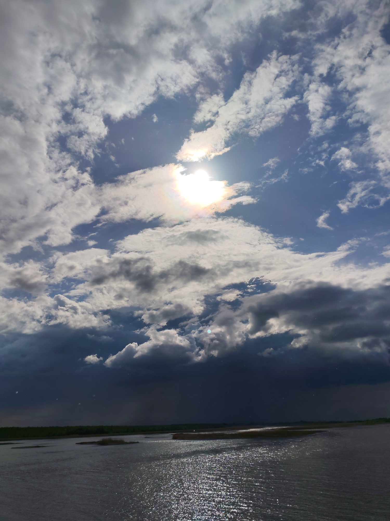 The image shows a body of water, likely a river or lake, under a dramatic sky. The upper portion of the sky is filled with a mix of white and gray clouds, with the sun visible through a break in the clouds, creating a bright halo effect. The lower part of the sky is dominated by dark, heavy storm clouds. The water surface reflects the sunlight, causing a shimmering effect. In the distance, there's a glimpse of land with some vegetation. The image likely shows a scene in Krokhino, Russia, although there are no specific landmarks visible to confirm the location definitively. There are no people present in the image. The focus is on the weather and its effect on the landscape.