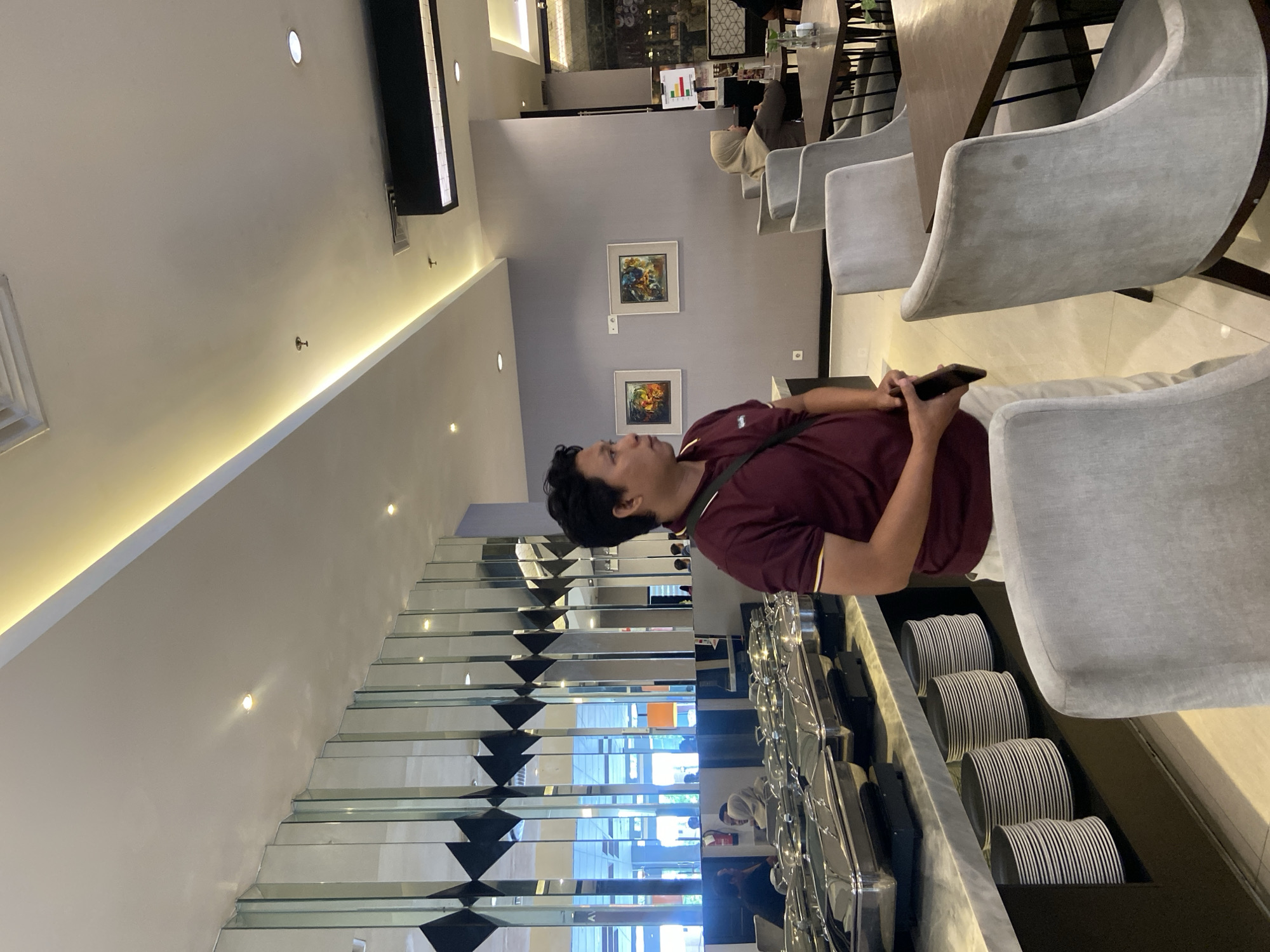 A man in a maroon shirt stands in a brightly lit interior, holding a phone in his hands. He is positioned next to a buffet setup featuring rows of stacked plates and covered serving dishes. A staircase with mirrored panels is visible in the background. Artwork hangs on the wall. The scene appears to be set in a restaurant, possibly within the city of Surabaya, Indonesia. Tables and chairs are seen in the periphery, with other people also present in the background.